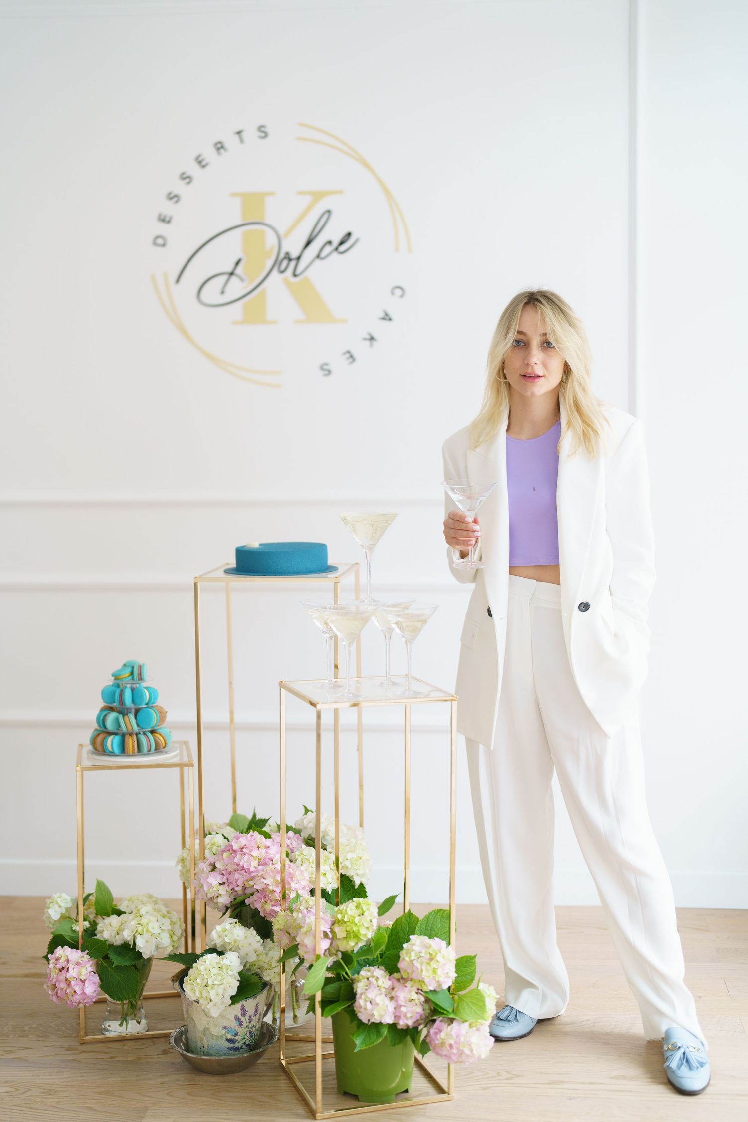 Blonde woman in white suit holding champagne glass at Dolce K Desserts Cakes shop with flowers and macarons display.