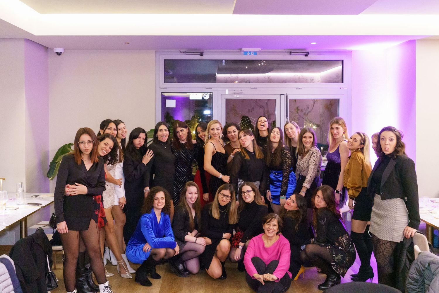 Group of women posing together at a purple-lit indoor event, smiling for a group photo.