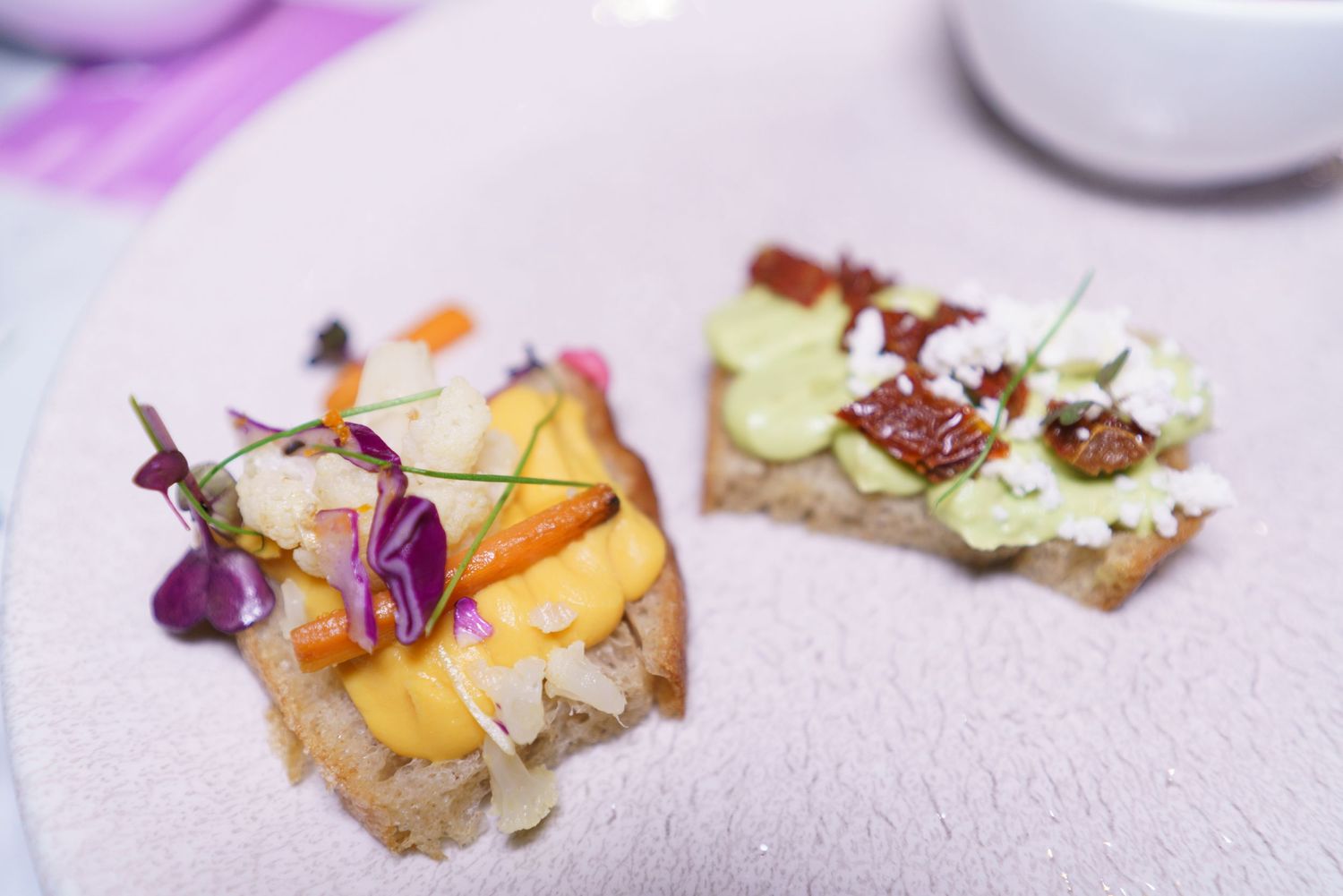 Two gourmet open-faced sandwiches on crispy bread topped with colorful vegetables, edible flowers, avocado, and sun-dried tomatoes.