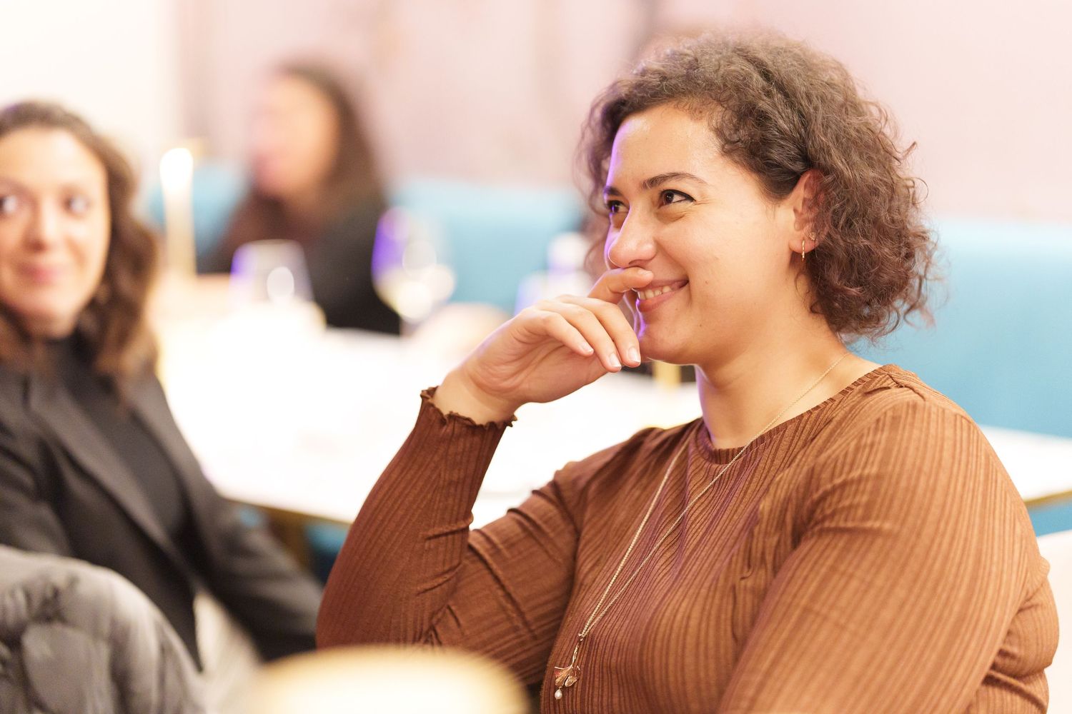 A smiling woman with curly hair in a brown top listens attentively at an indoor event.