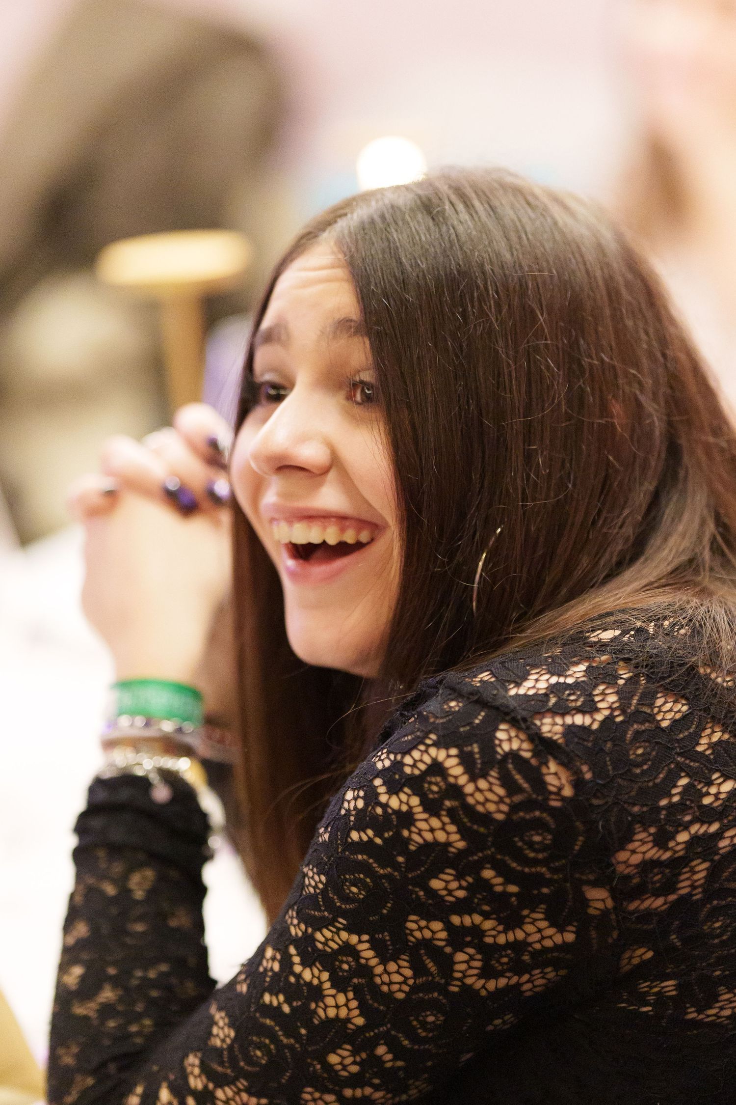 A young woman with long brown hair laughs joyfully while holding a phone, wearing a black lace top and green bracelet.