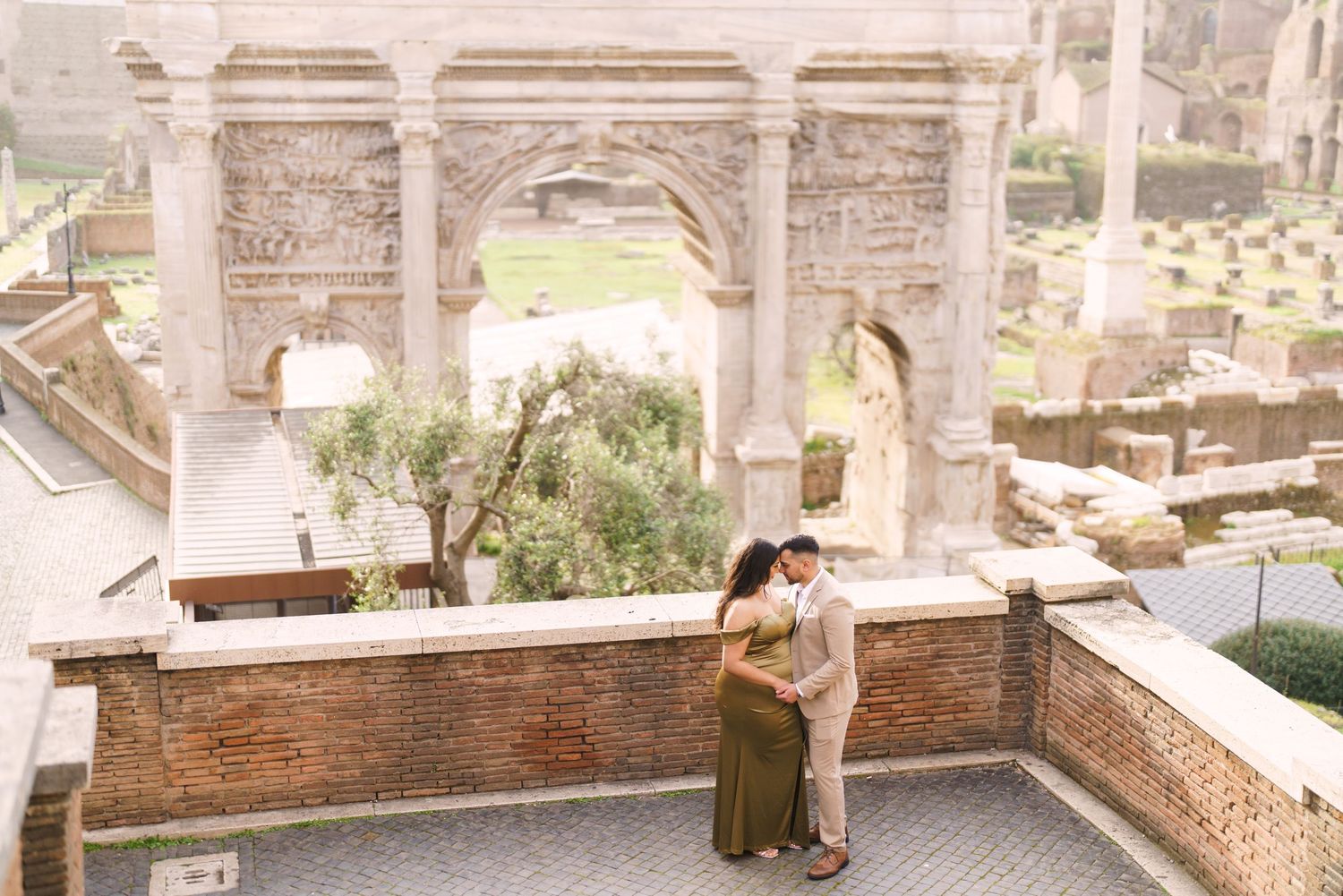 Couple embracing near ancient Roman arch ruins at the Roman Forum, woman in green dress, man in beige suit.