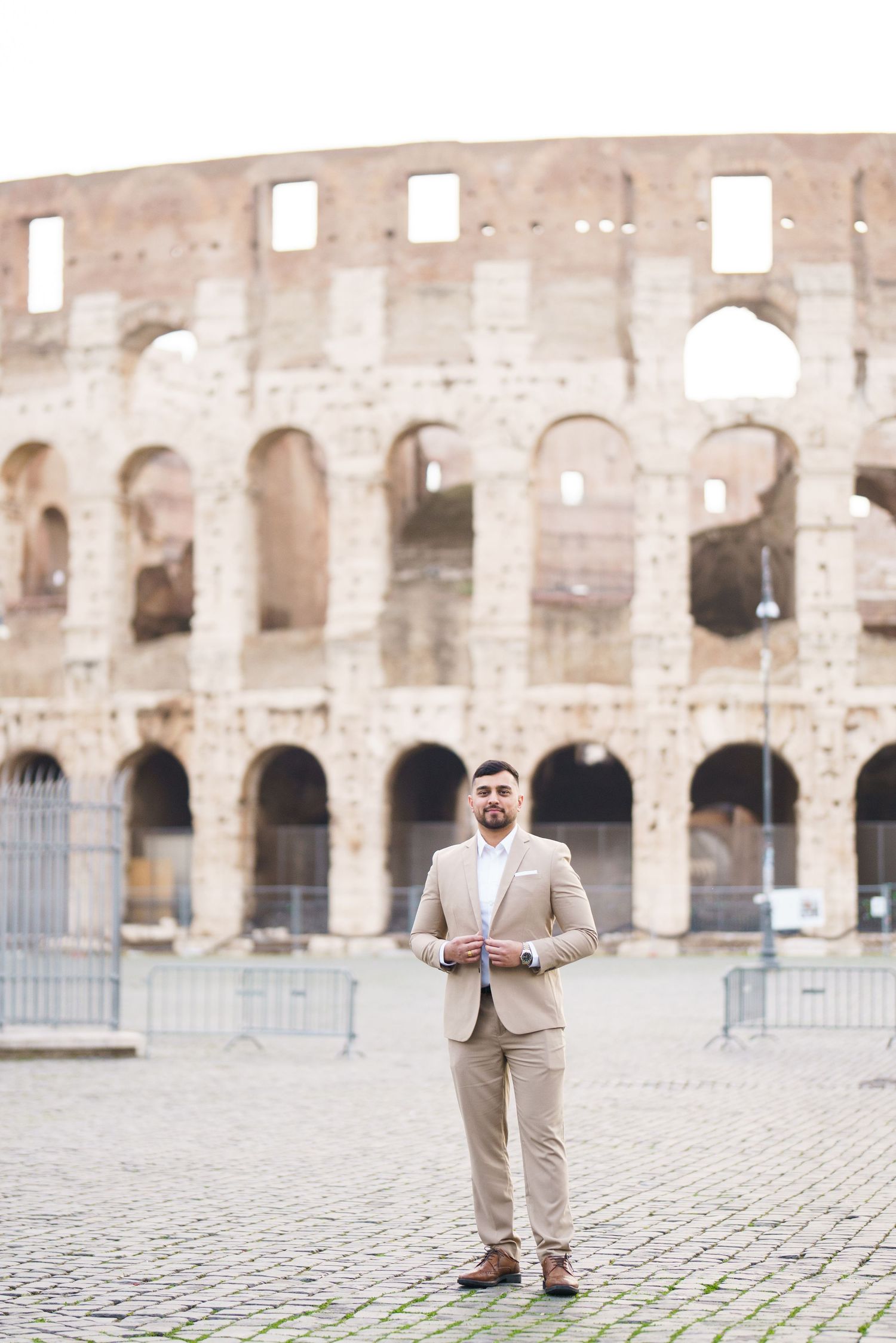 A well-dressed man in a beige suit stands confidently in front of the ancient Roman Colosseum in Rome, Italy.