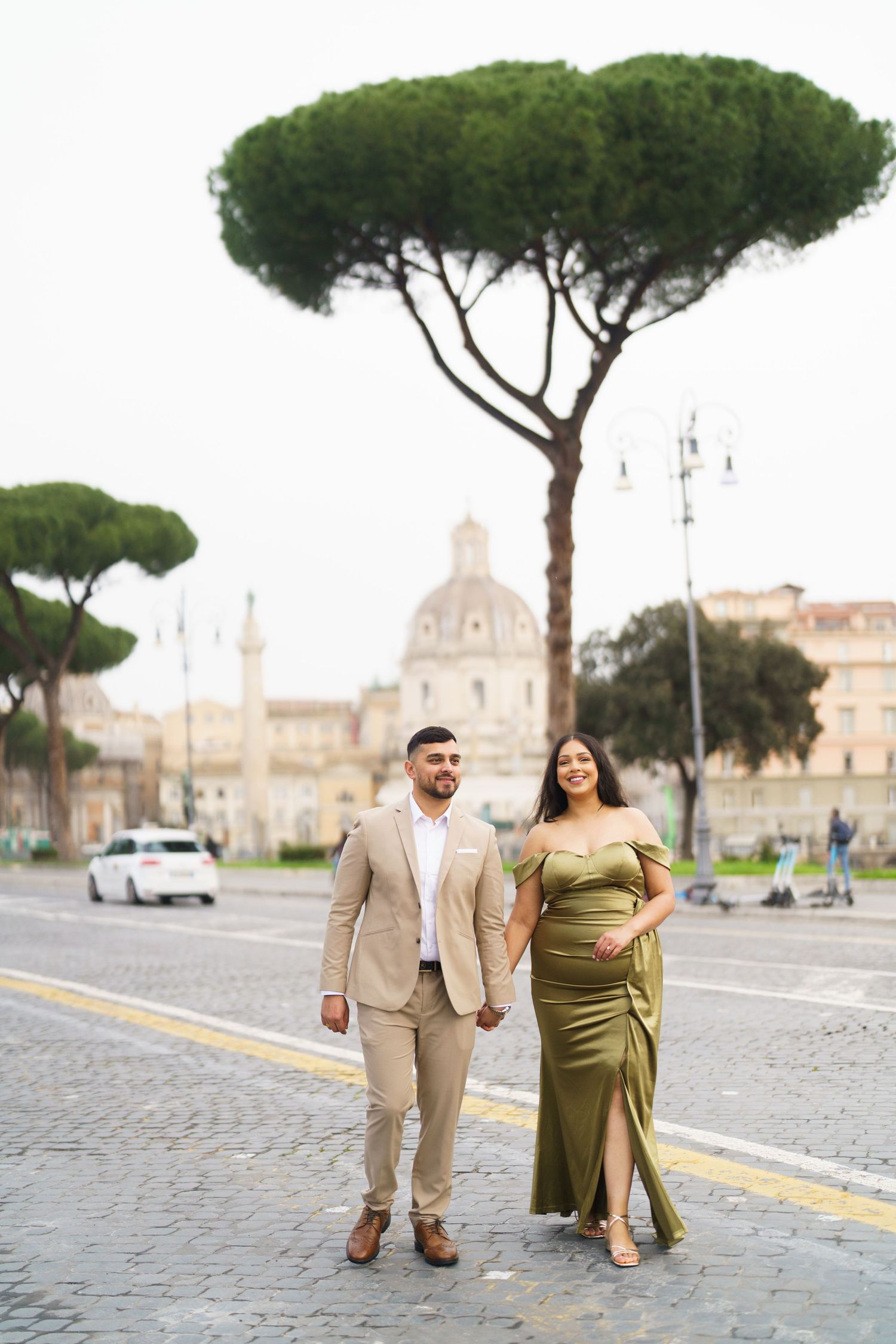 Elegant couple holding hands walking on cobblestone street in Rome, woman in green gown, man in beige suit, church backdrop.