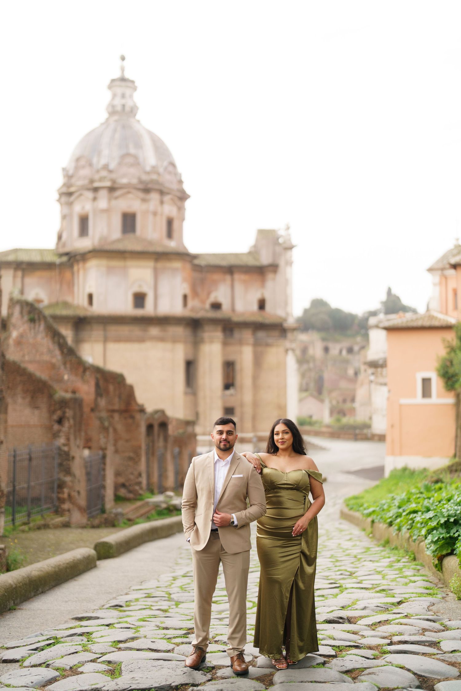 Couple posing on cobblestone path in Rome, woman in green gown, man in tan suit, historic church dome behind them.