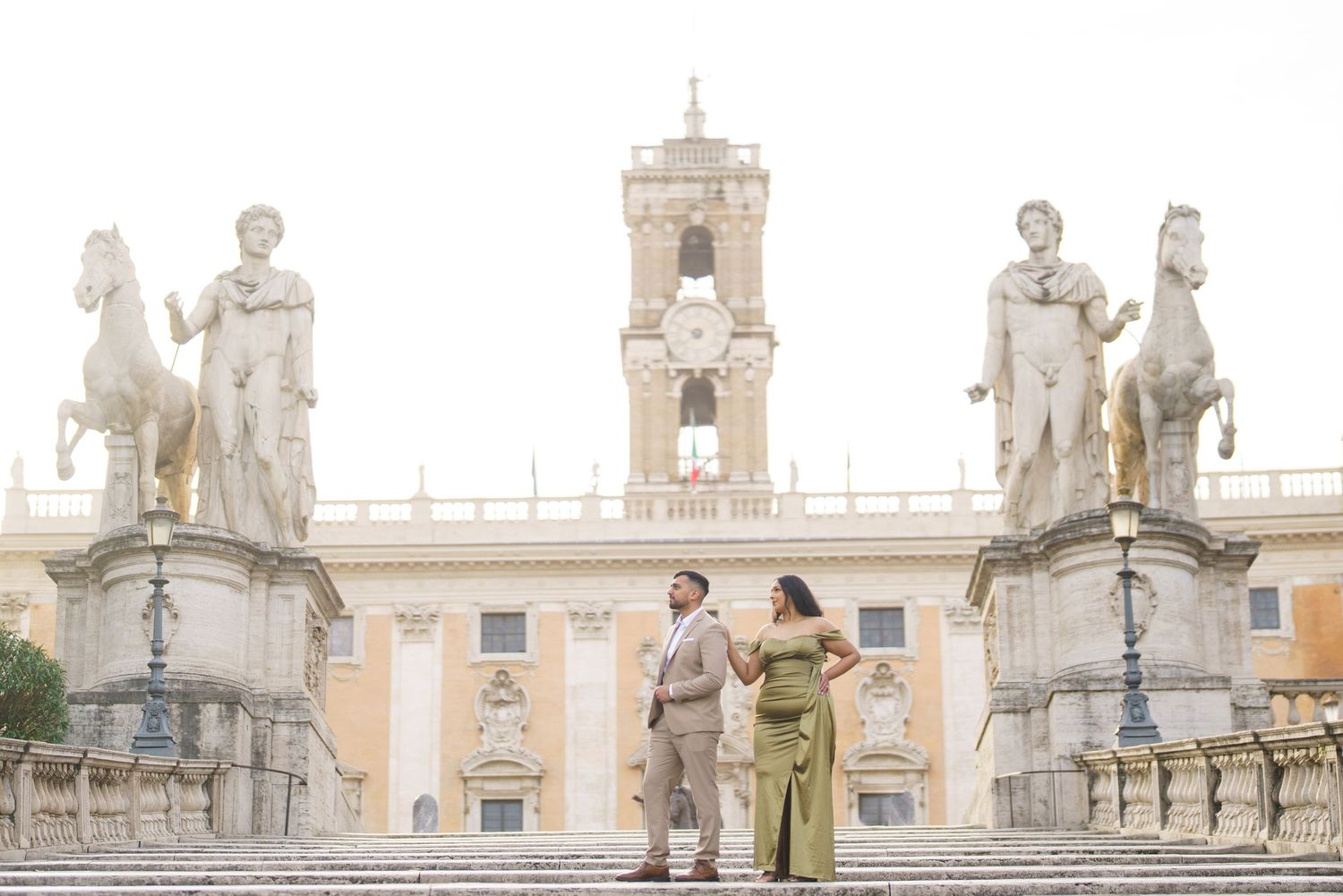 Couple posing on steps of Capitoline Hill in Rome, flanked by classical statues and a historic clock tower.