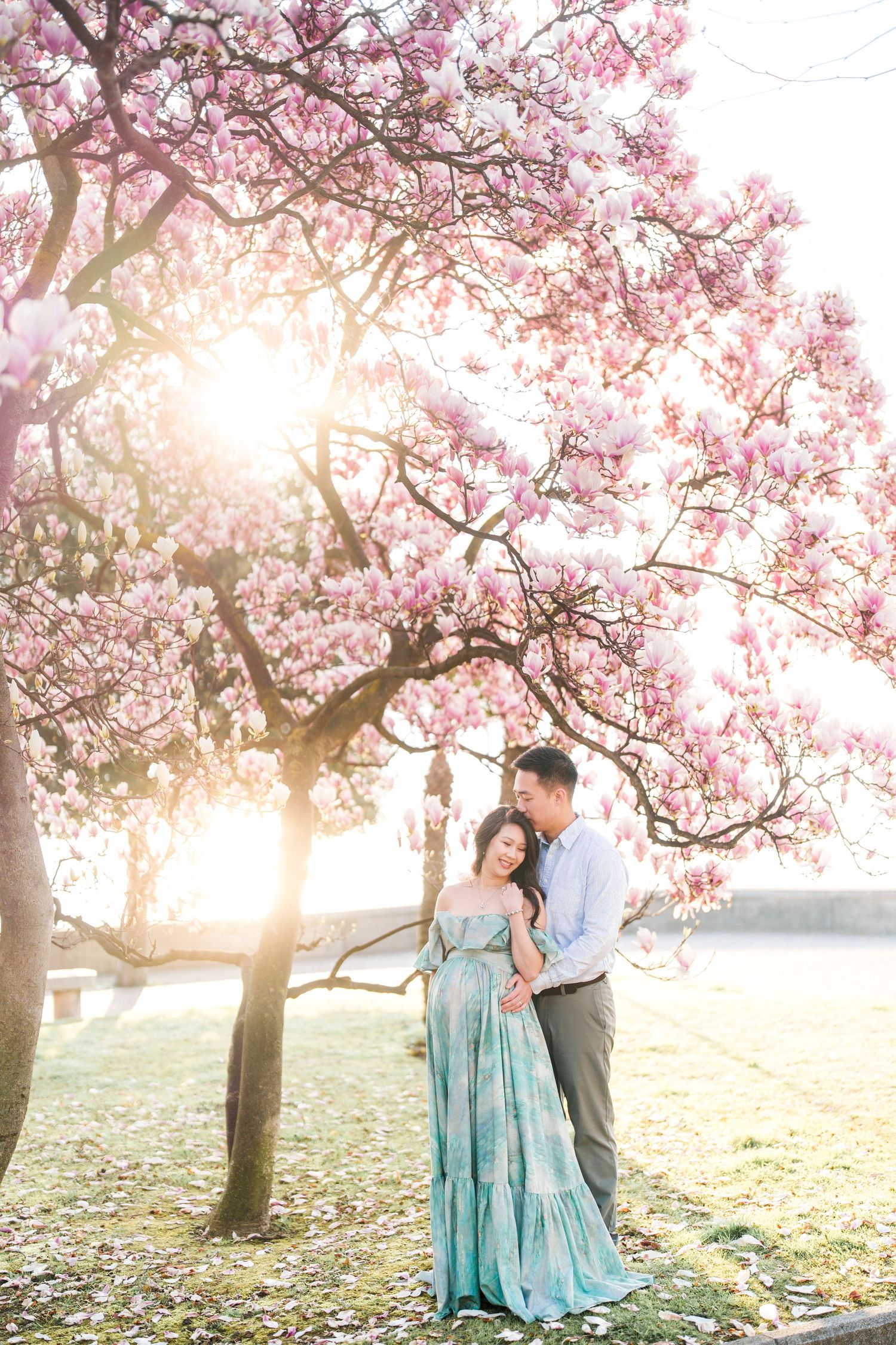 Couple embracing under blooming pink magnolia tree at golden hour in a romantic outdoor engagement photo.