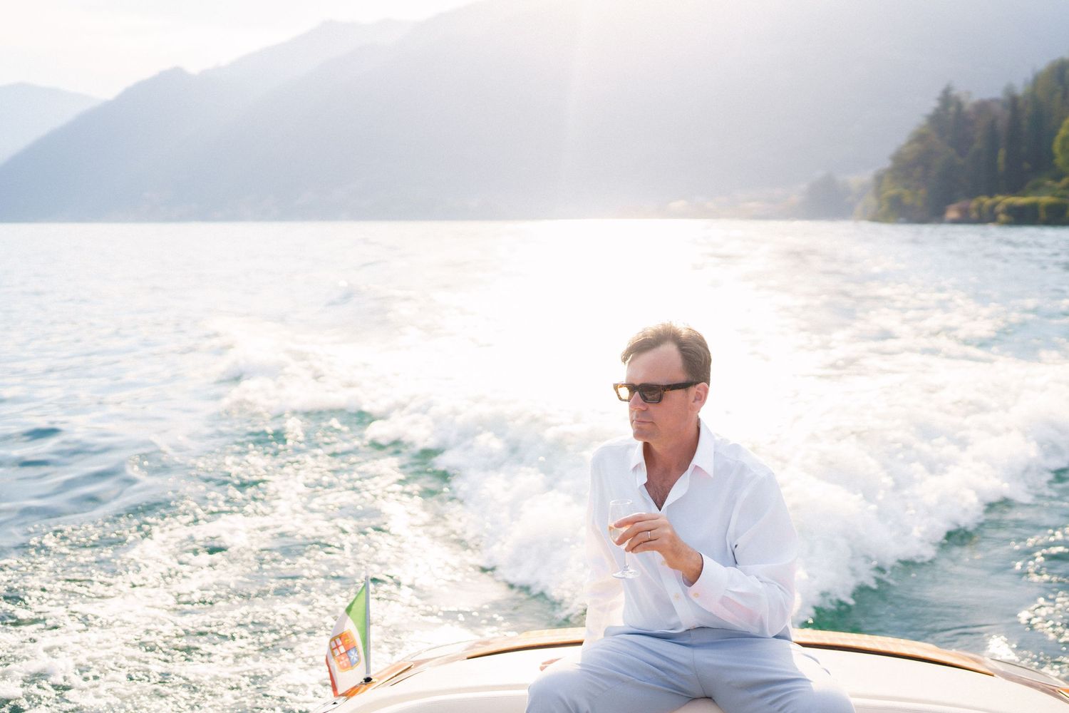 Man in white shirt sitting on a boat on a serene mountain lake, enjoying the sunny day with wake behind him.