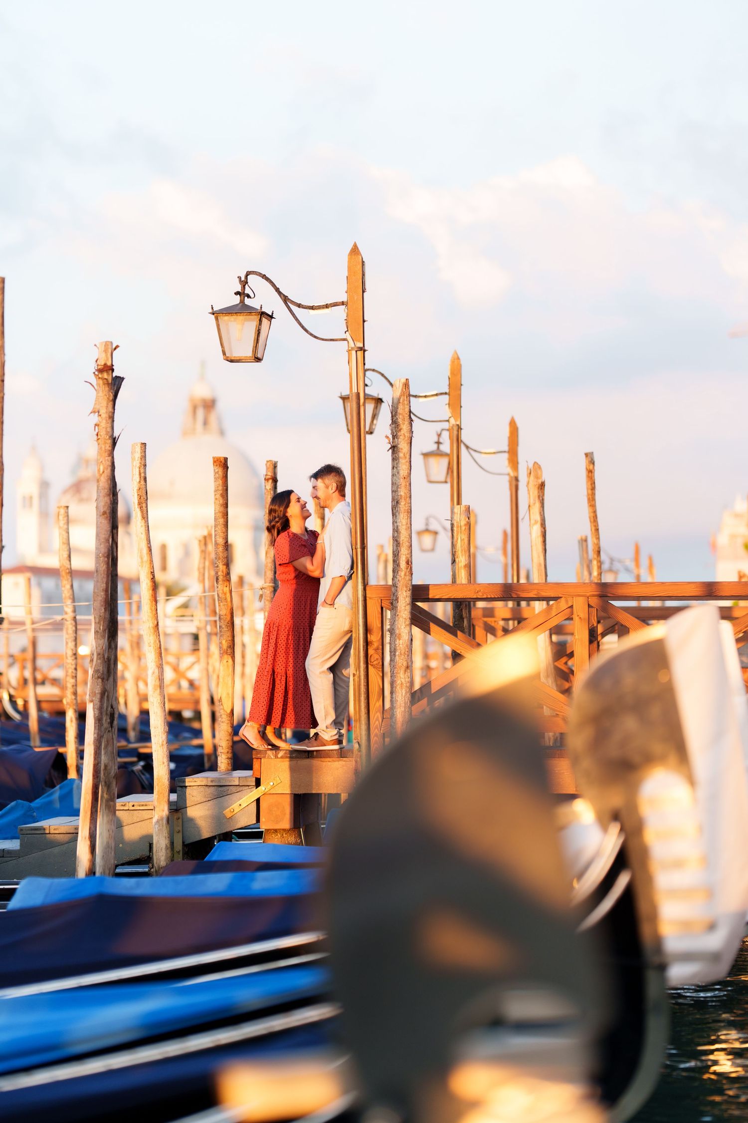 Romantic couple embracing on Venice gondola dock at sunset with classic lamp posts and Santa Maria della Salute in background.
