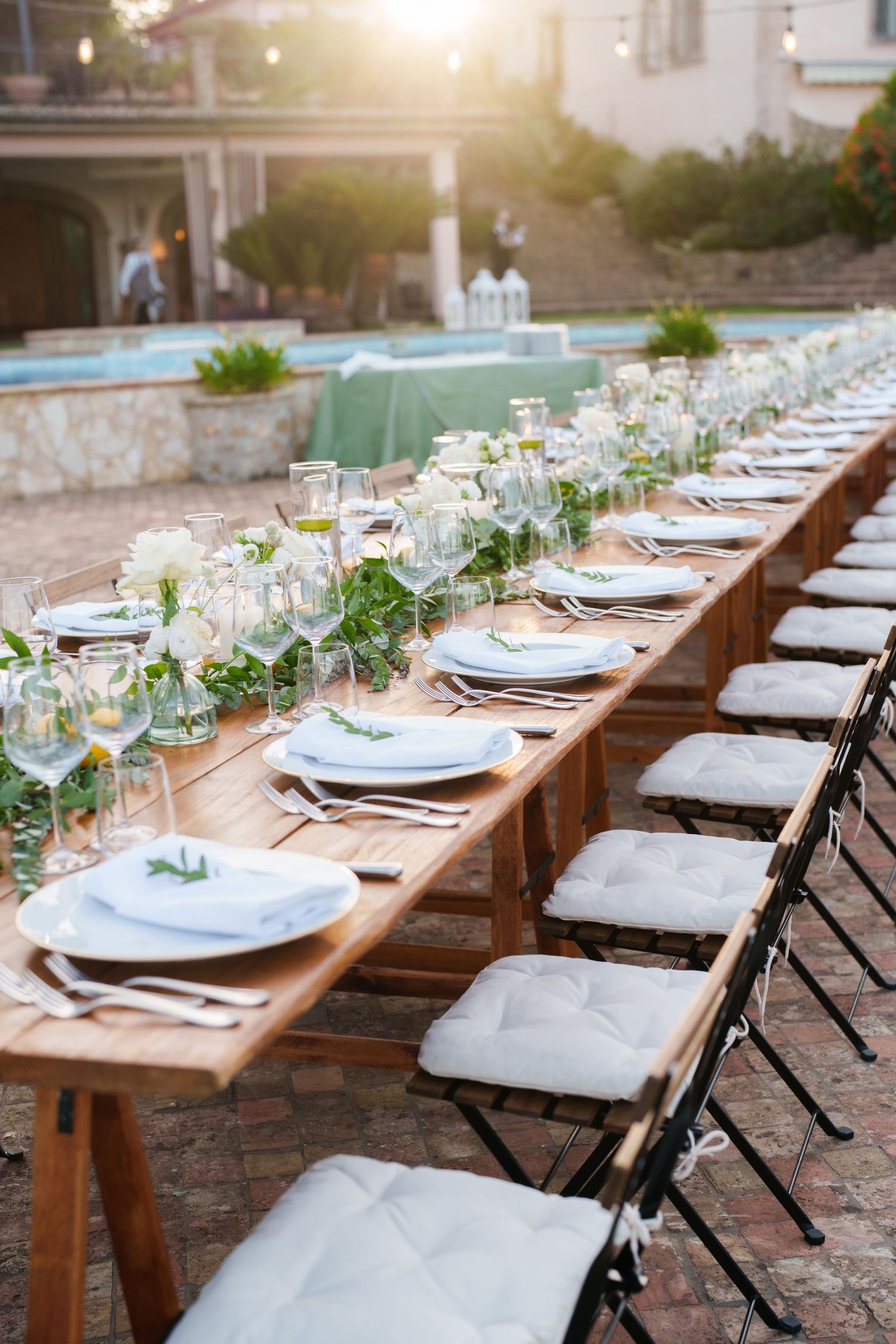 Elegant outdoor wedding reception with long wooden table, white place settings, floral greenery, and cushioned chairs at golden hour.