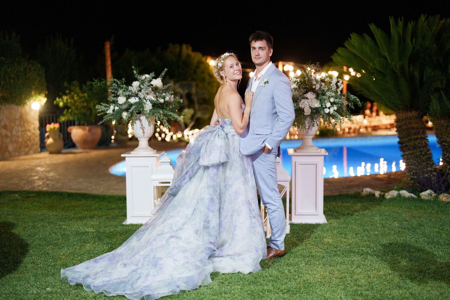 Bride in blue floral gown and groom in gray suit pose elegantly outdoors at night wedding reception.