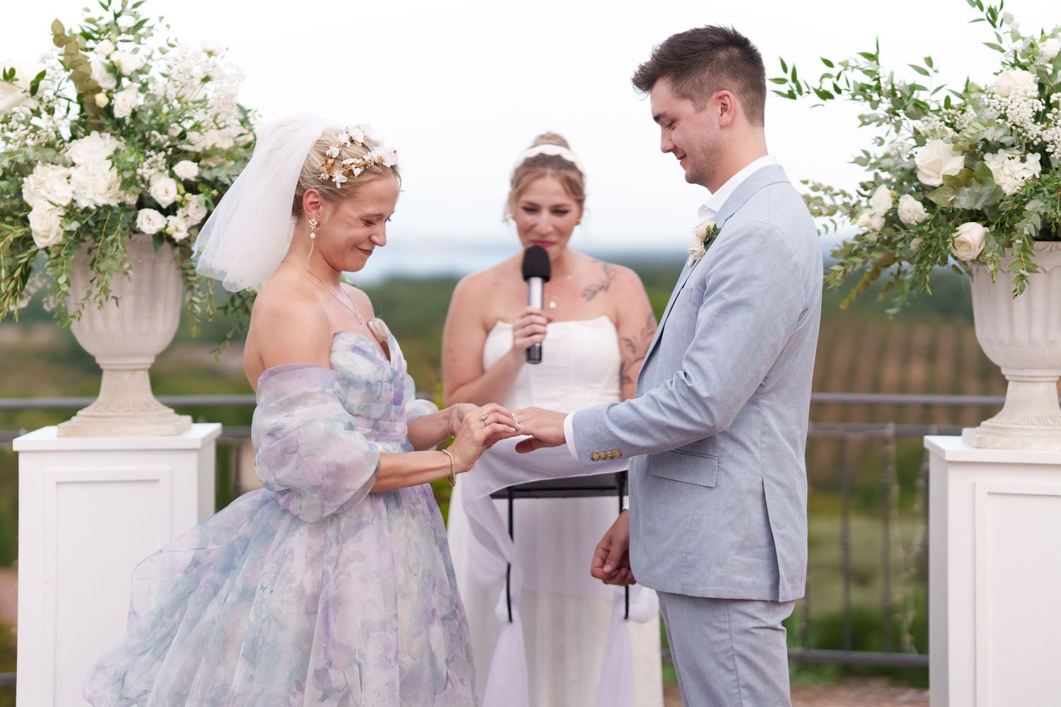 Bride in blue floral gown exchanges rings with groom in gray suit during outdoor wedding ceremony with white floral arch.