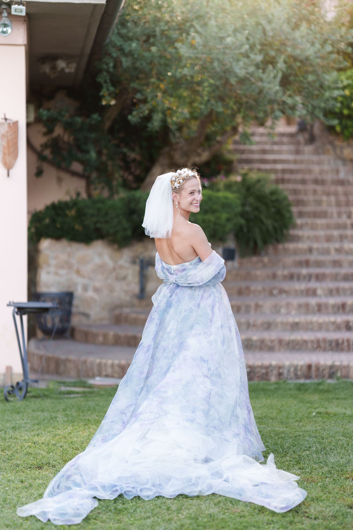 Bride in elegant blue floral off-shoulder gown with veil and floral headpiece poses on green lawn near stone steps.