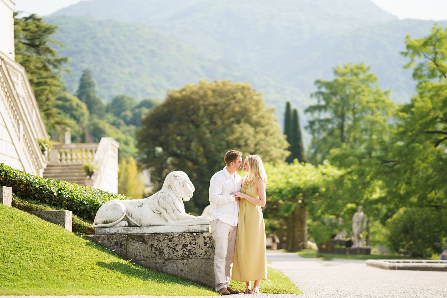 Couple kissing in lush garden with stone sphinx sculpture and mountain backdrop at an Italian villa.