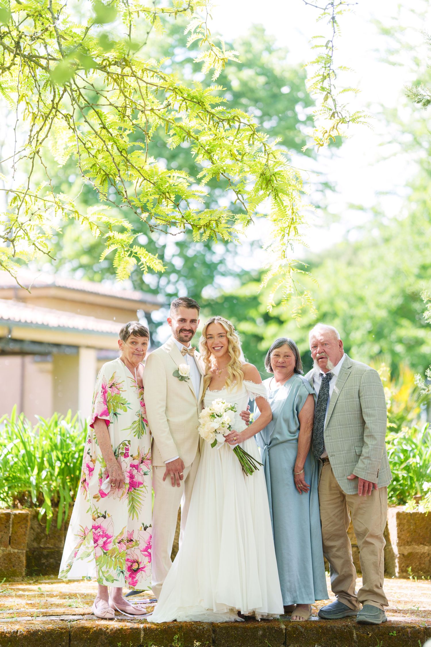 Wedding couple poses with family outdoors under lush green trees, bride holding white bouquet in elegant off-shoulder gown.