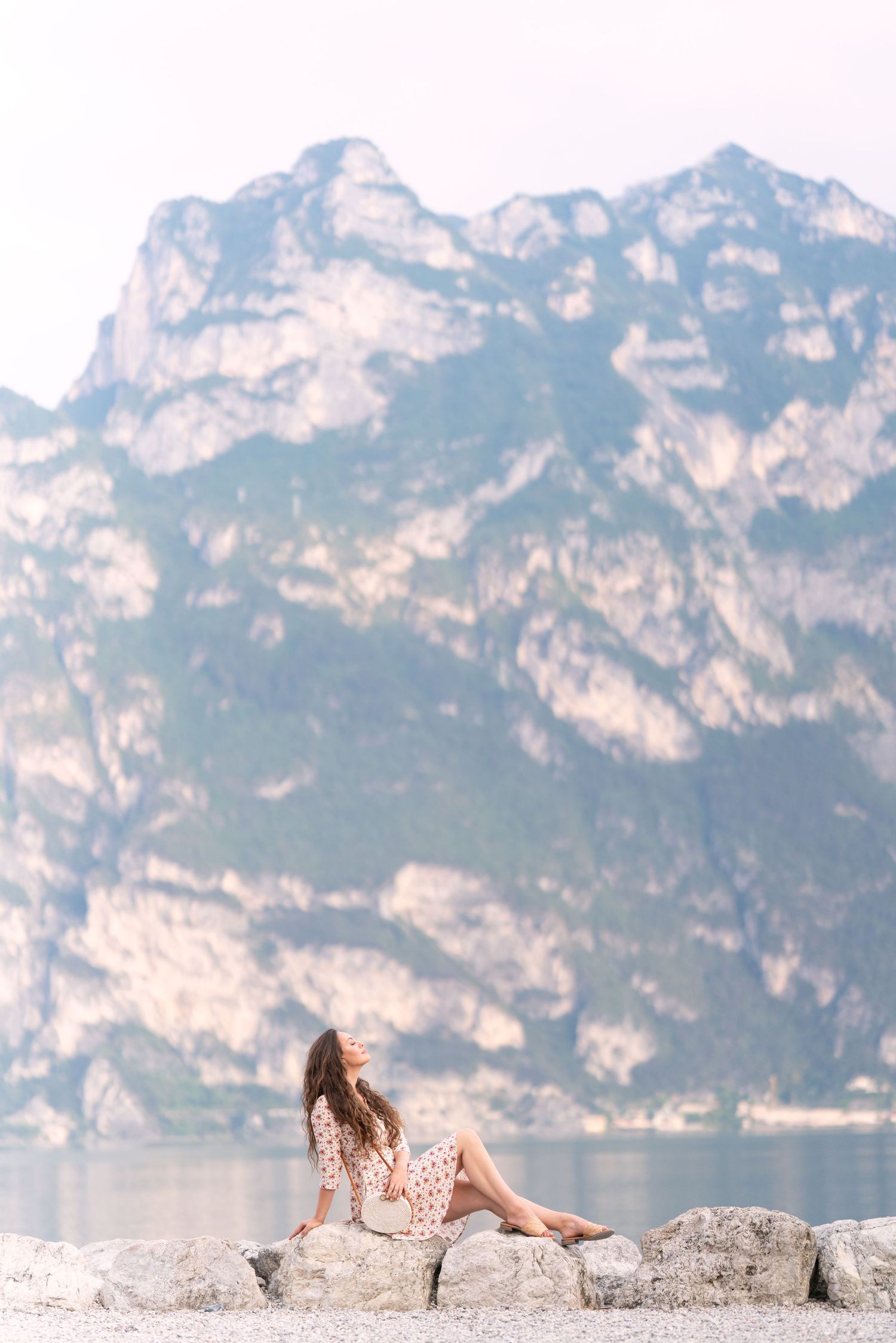 Woman in floral dress sitting on rocks by a mountain lake with dramatic rocky peaks in the background.