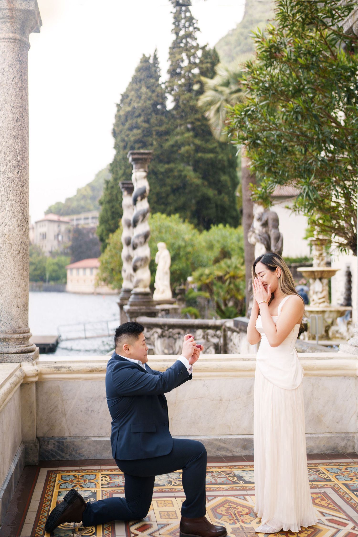 Man proposes on one knee to woman in white dress on ornate terrace overlooking Lake Como, Italy.