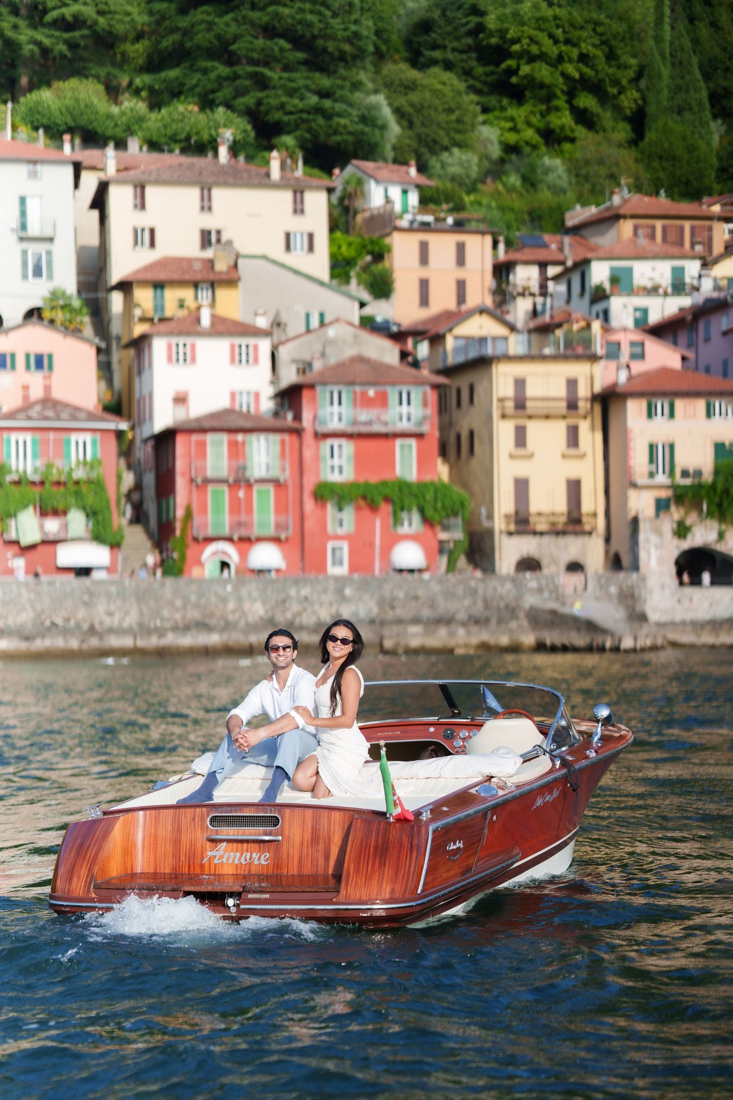 Couple riding a classic wooden speedboat on Lake Como with colorful Italian village buildings in the background.