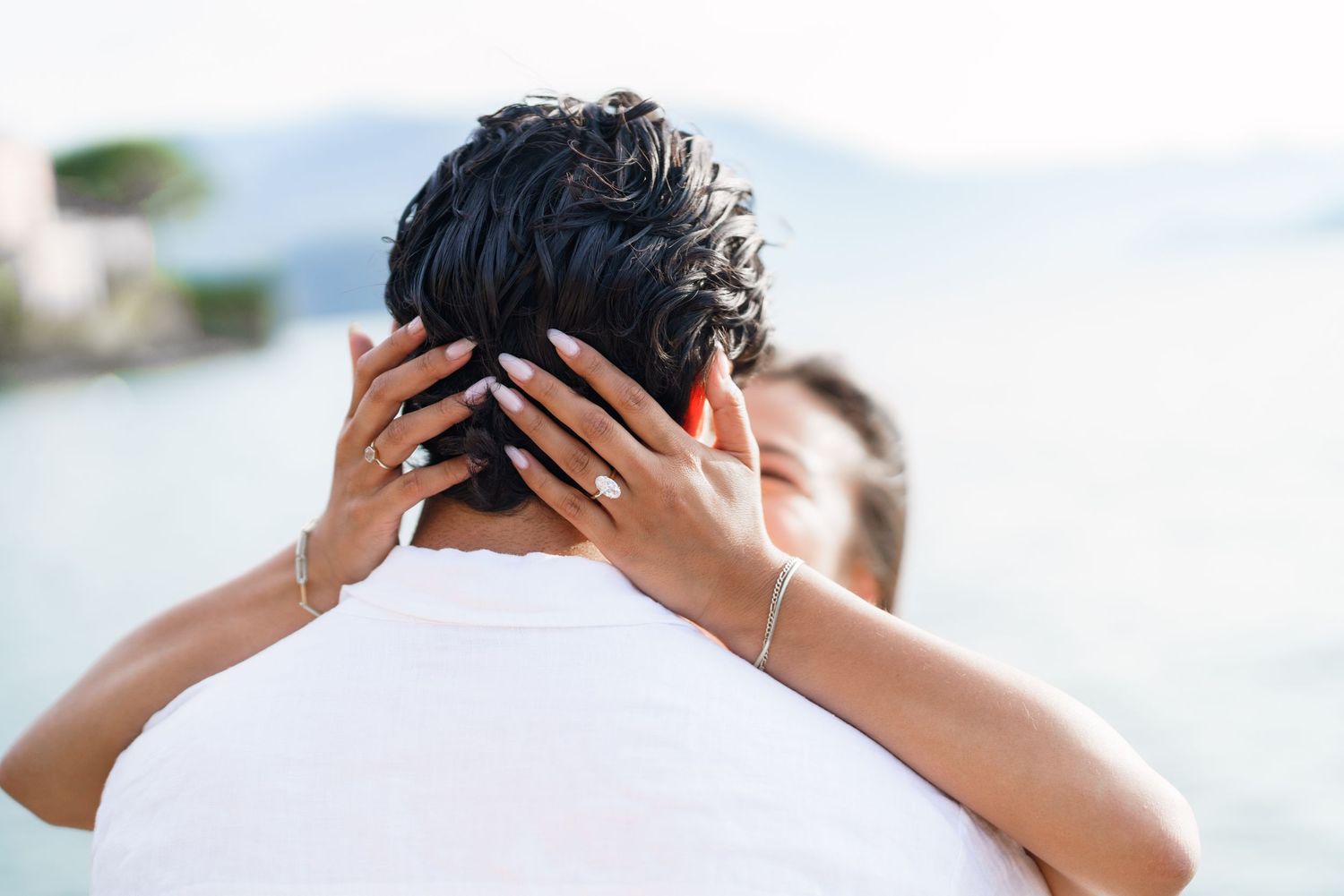 Couple embracing outdoors near water, woman with ring and bracelet holding man's head in romantic moment.