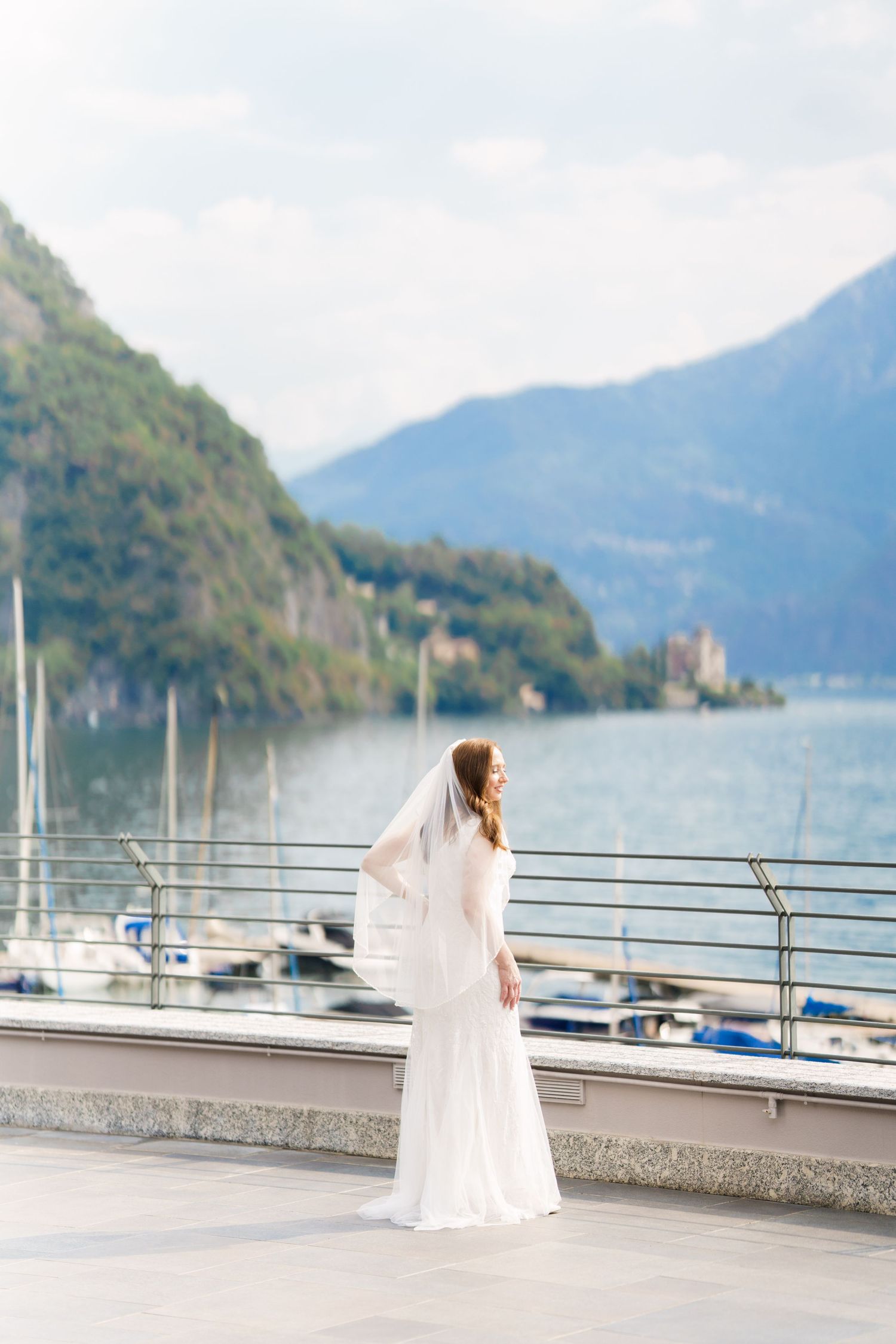 Bride in white wedding dress and veil stands on lakeside promenade with mountain backdrop and marina views.