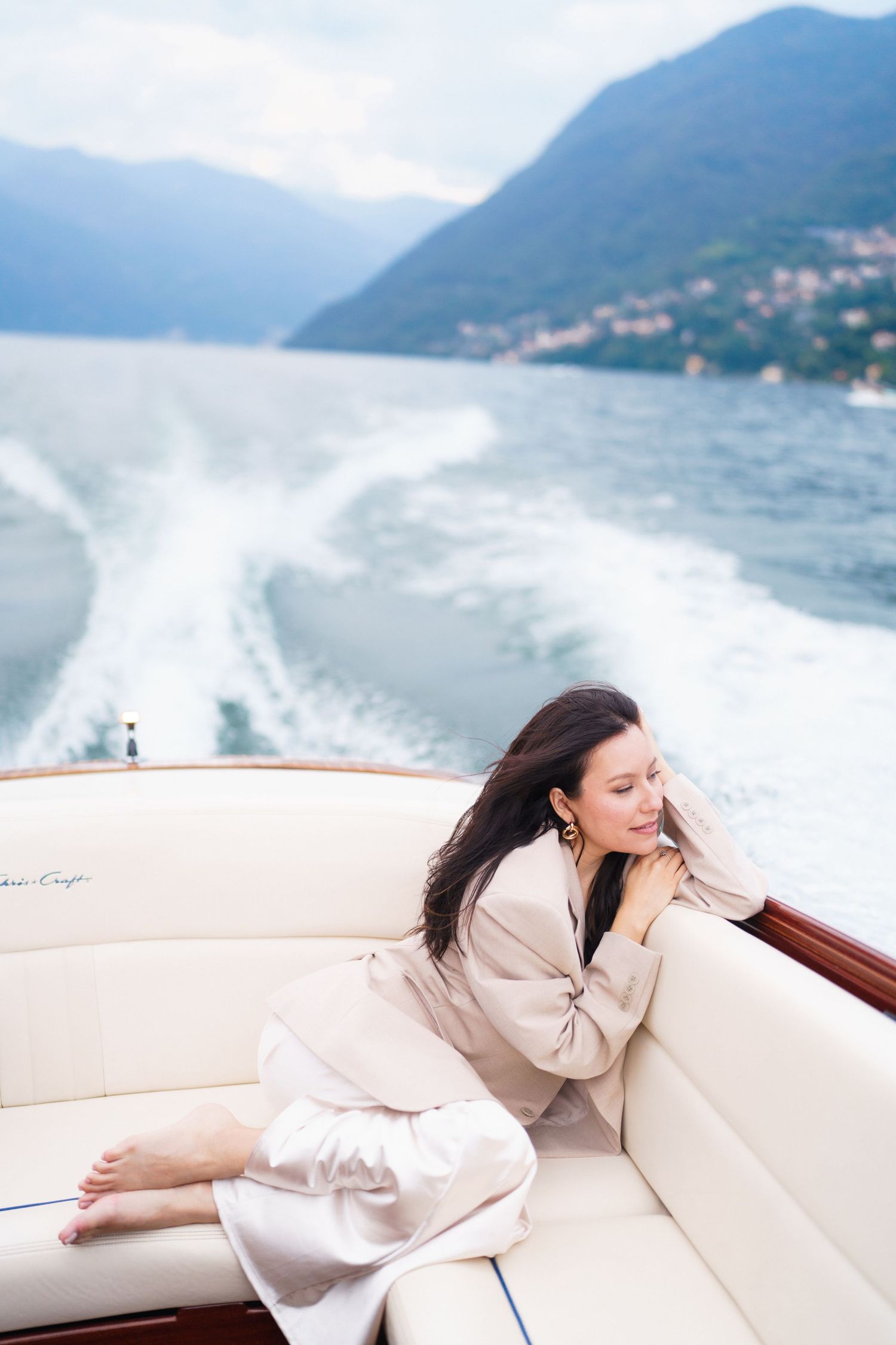 A woman in beige loungewear relaxes on a luxury boat with scenic mountain views over Lake Como in the background.
