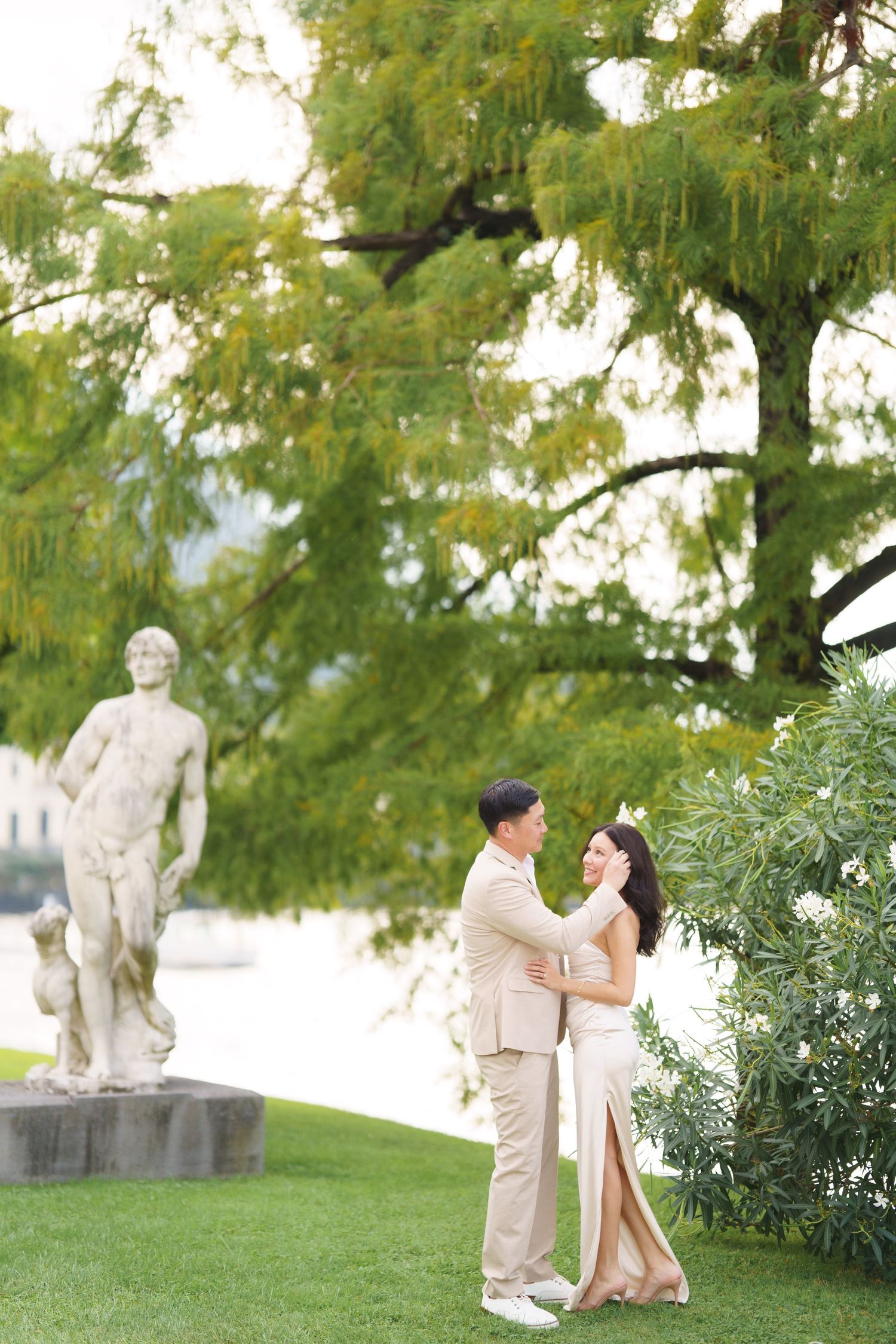 A couple shares a romantic moment in a garden setting with stone sculptures and lush greenery in the background.