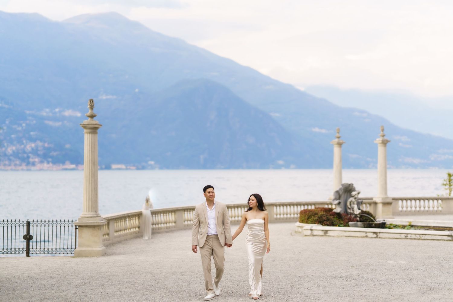 A couple walks along a scenic balcony overlooking Lake Como in Italy with ornate stone columns and mountain views.