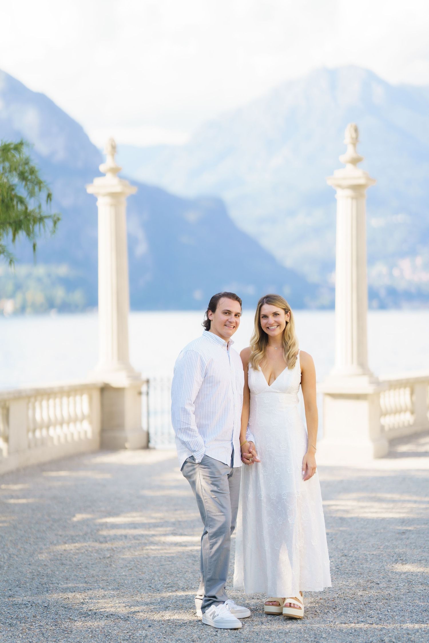 A couple in white attire poses together on a scenic terrace overlooking mountain views in Lake Como, Italy.