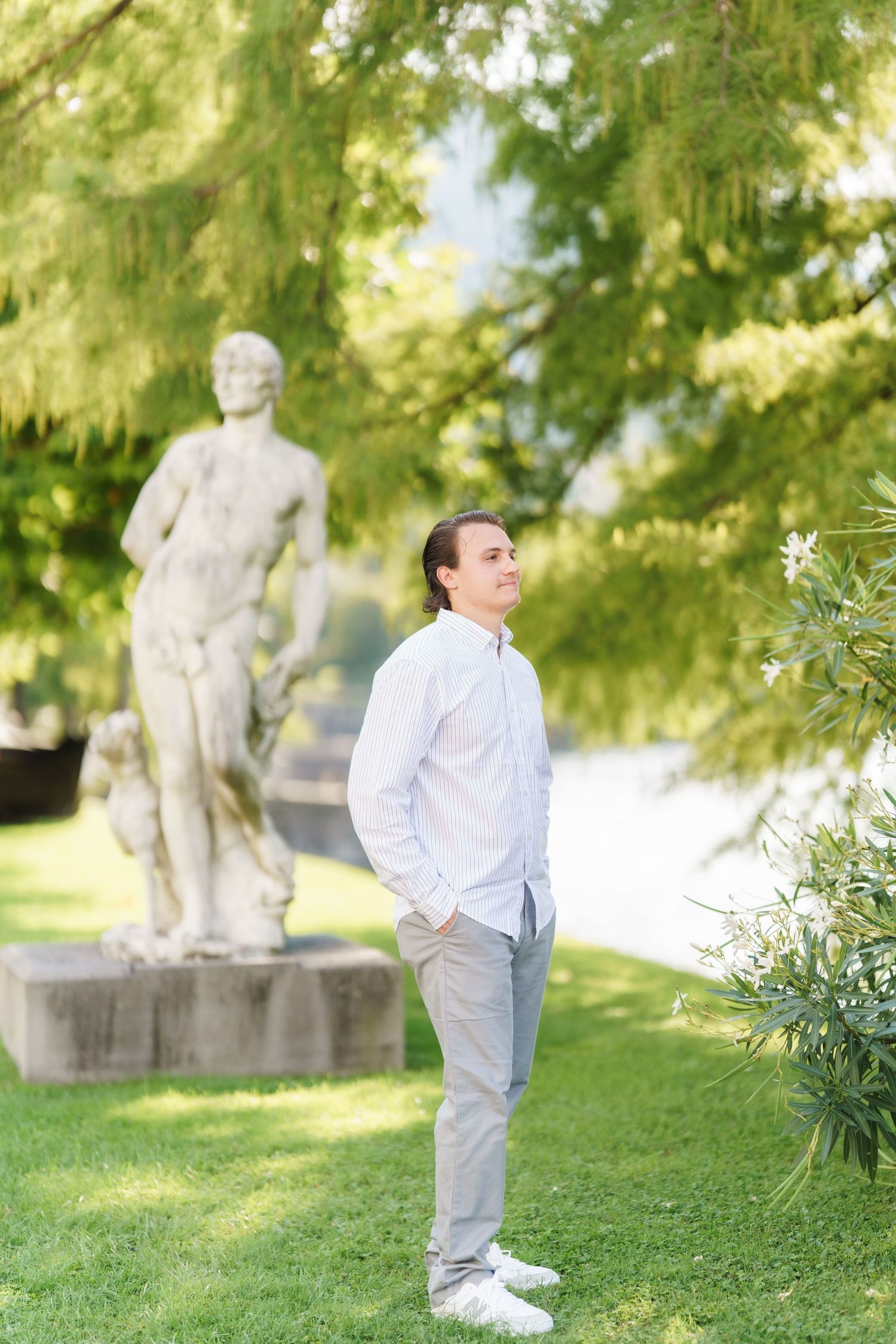 Young boy in white shirt and gray pants stands near garden statues in a sunlit park setting.