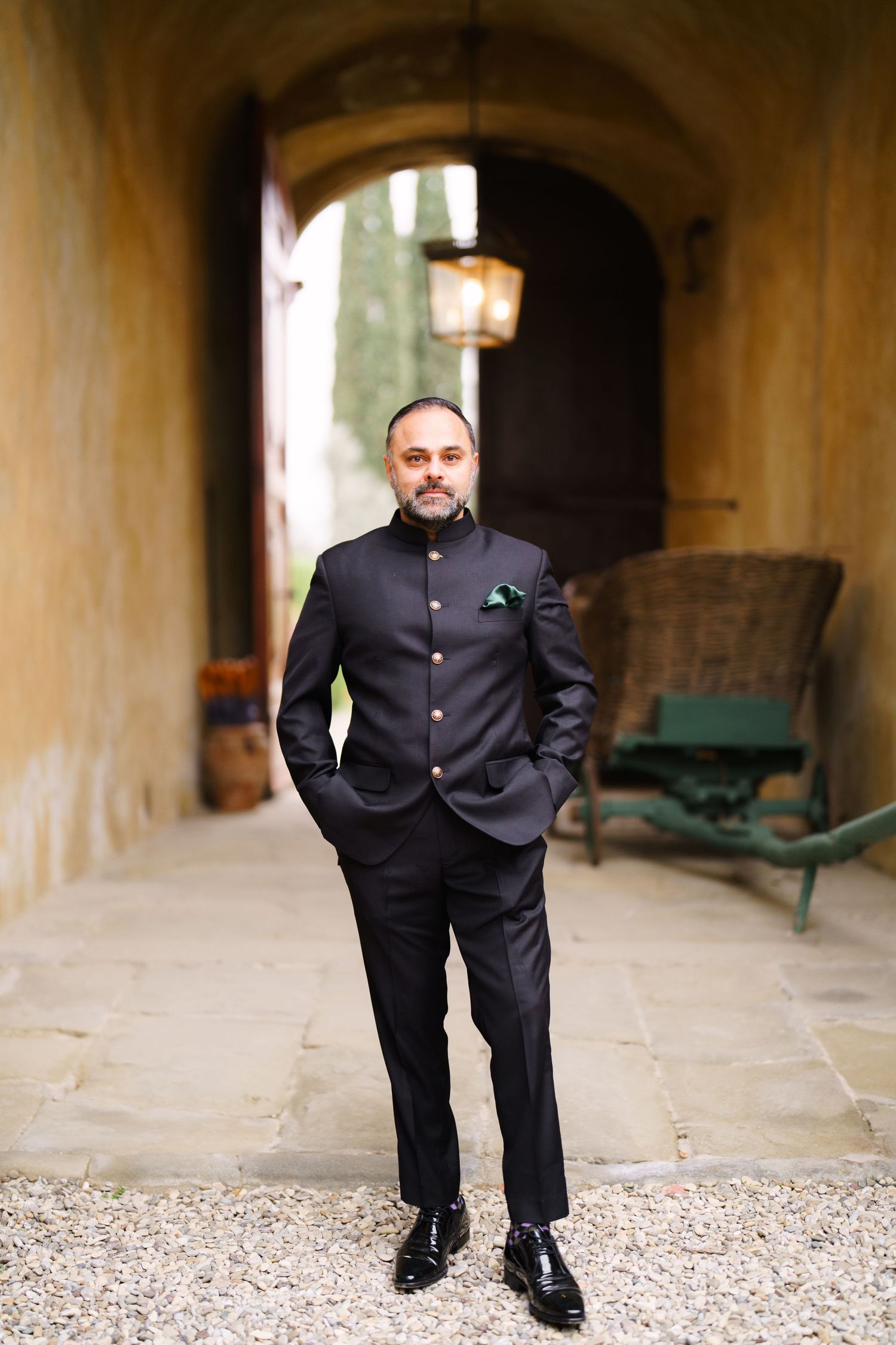 Man in formal black Nehru suit poses confidently under stone archway.