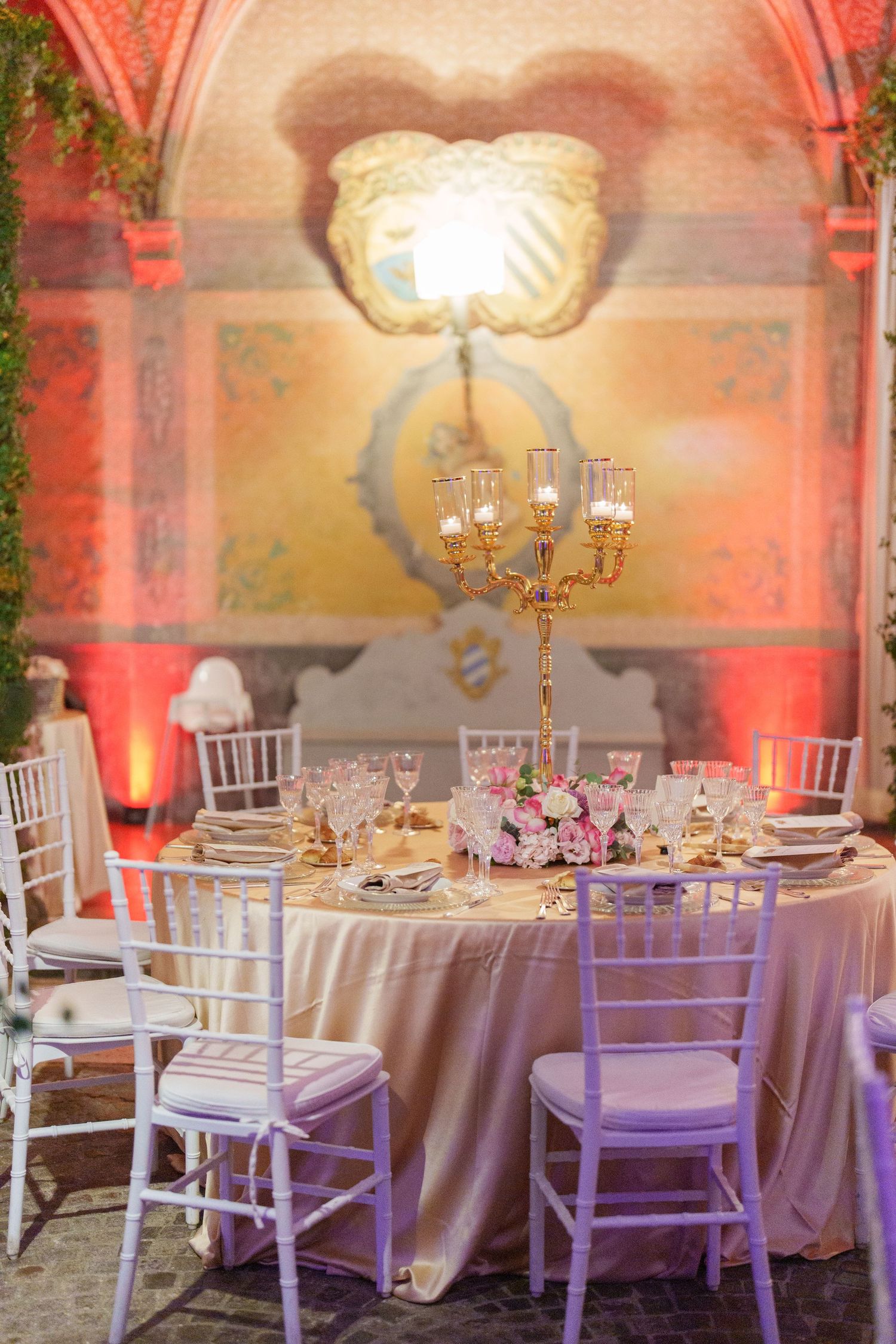 Formal dining setup with white chairs and tables in an ornate room with coral-colored accent lighting.