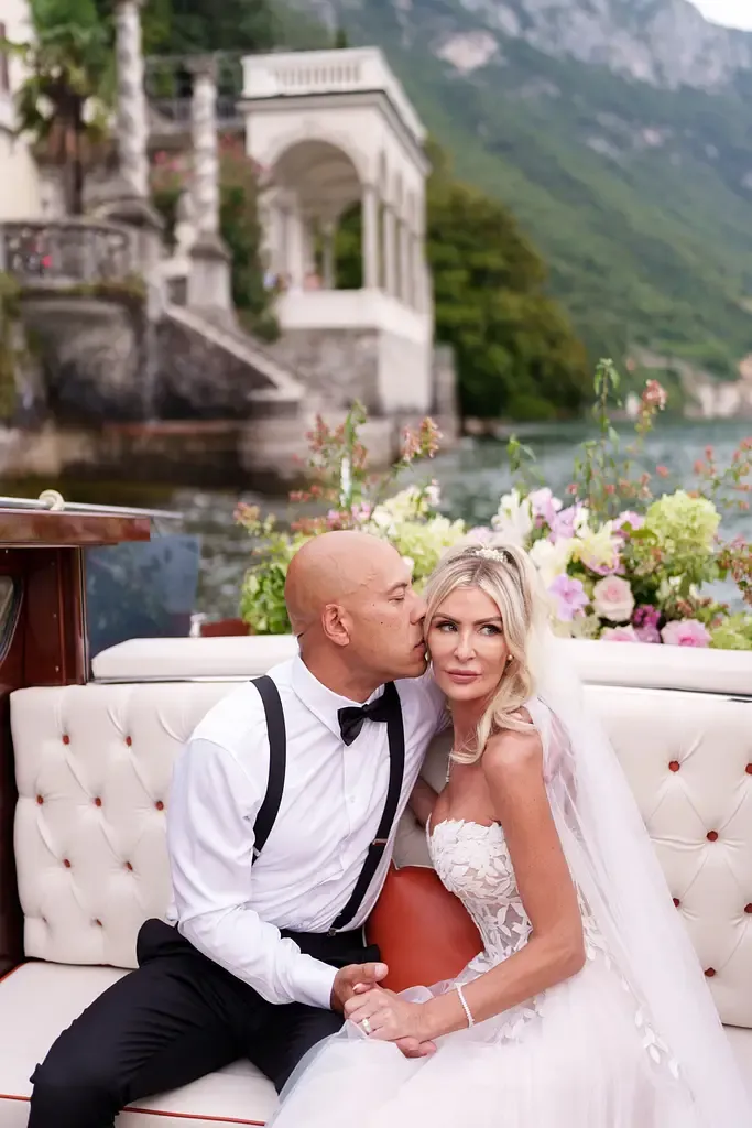 Romantic wedding couple lounges on elegant cream sofa with mountain lake backdrop at luxurious Italian villa.
