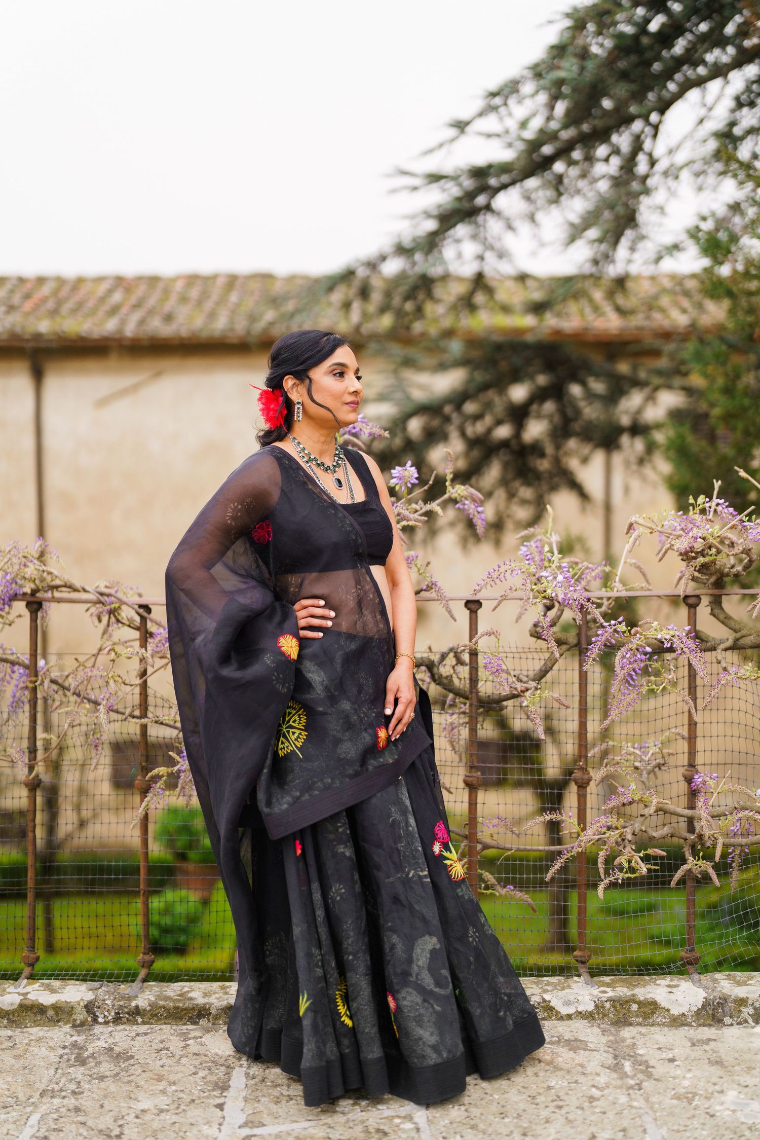 Person in elegant black dress posing by stone wall with blooming trees in background.