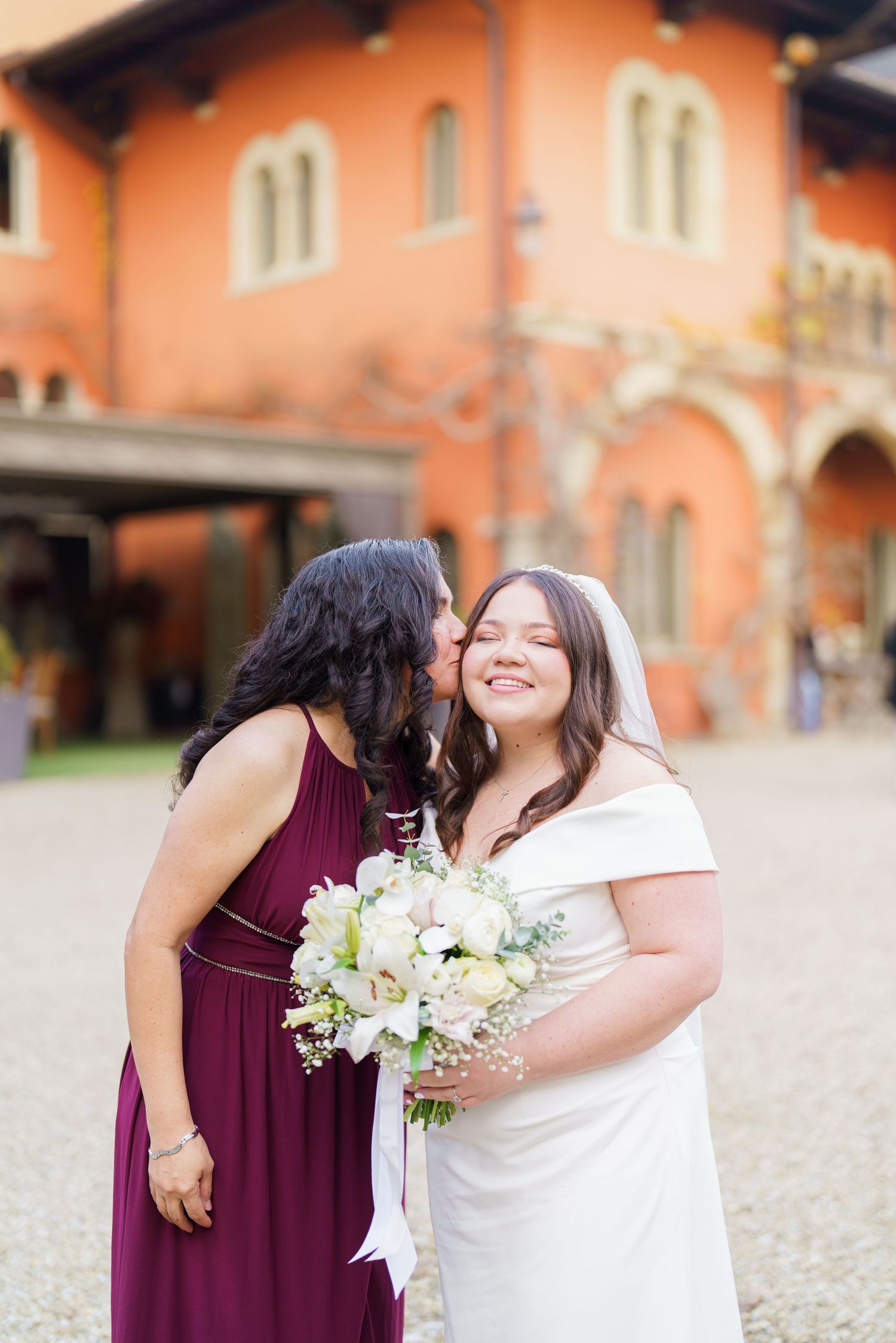 Two people share a joyful moment in burgundy and white dresses while holding a white floral bouquet.