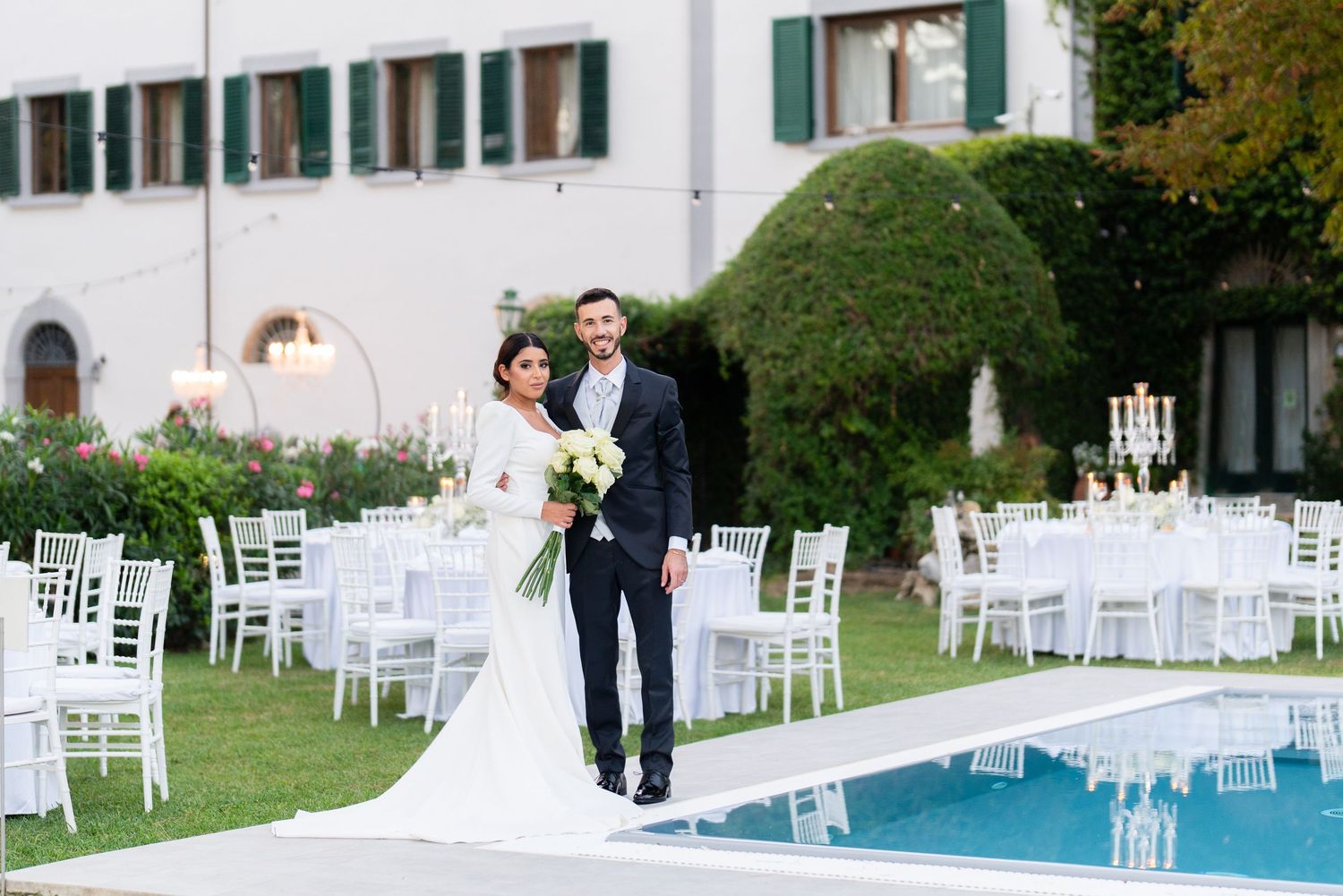 Elegant outdoor wedding reception setup with white tables and chairs around swimming pool.