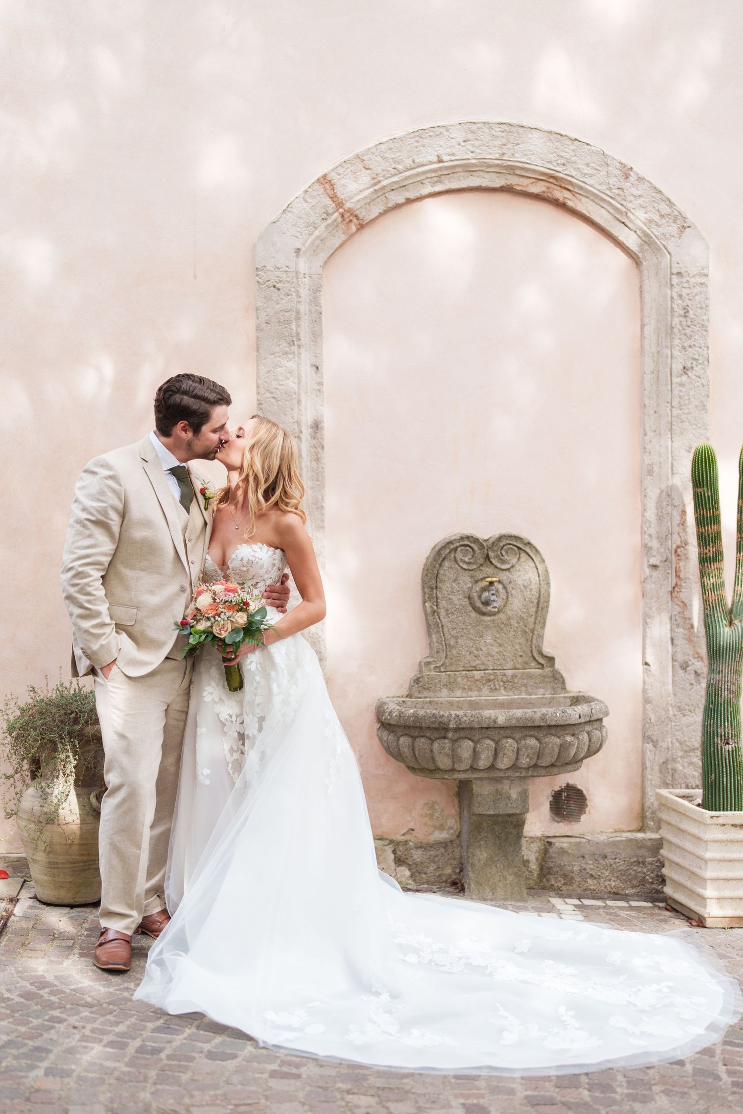 A romantic wedding couple shares a tender moment against a stone archway with an ornate fountain in the background.
