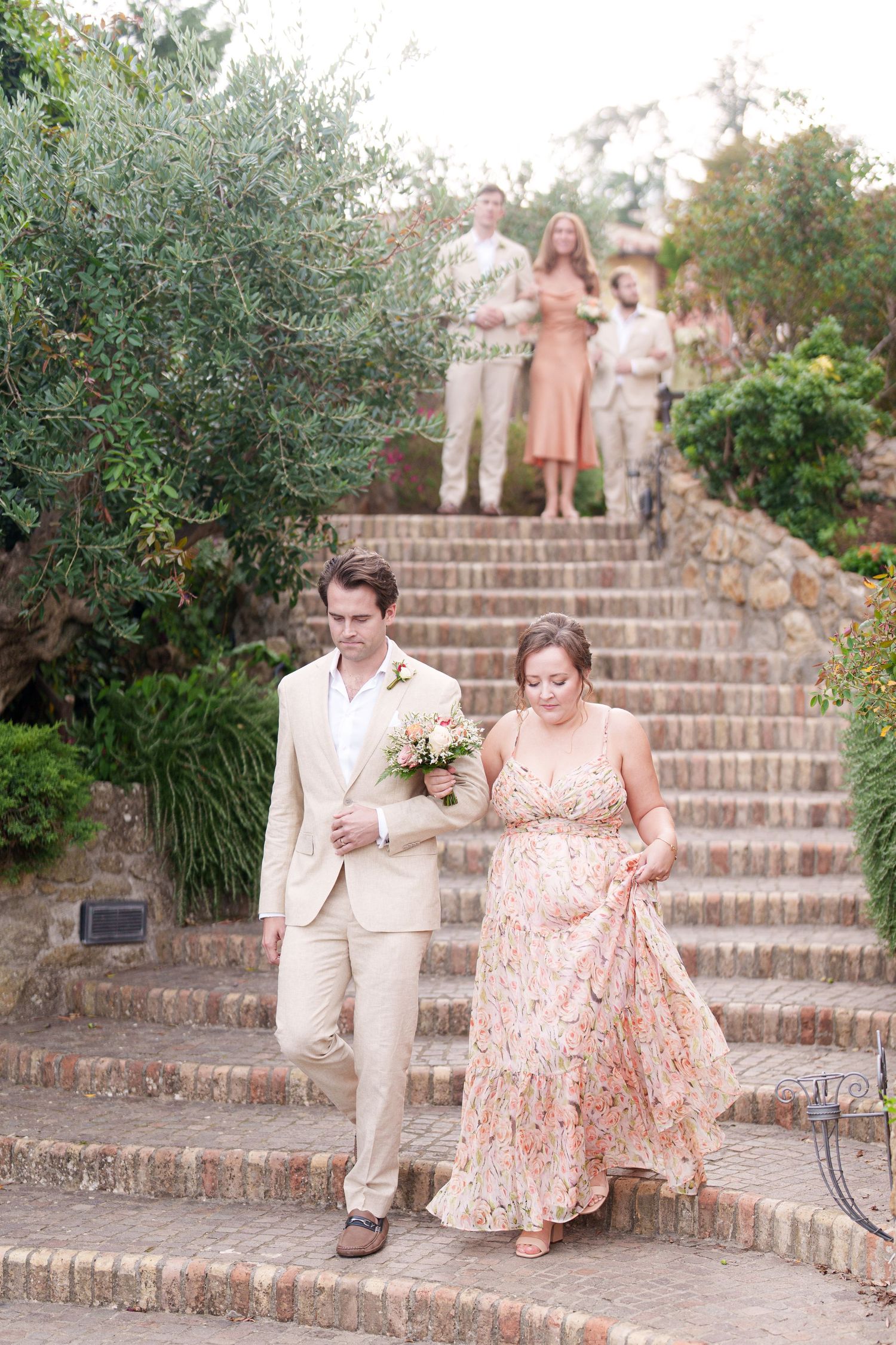 Couple in light-colored attire walking down rustic brick stairs during a celebration.