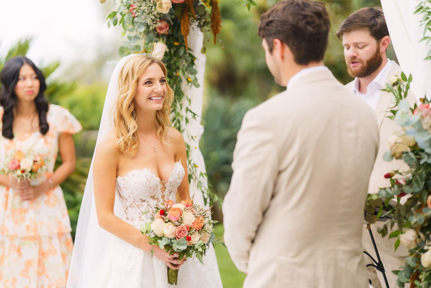 An intimate wedding ceremony moment under a floral arch with bridesmaids watching.