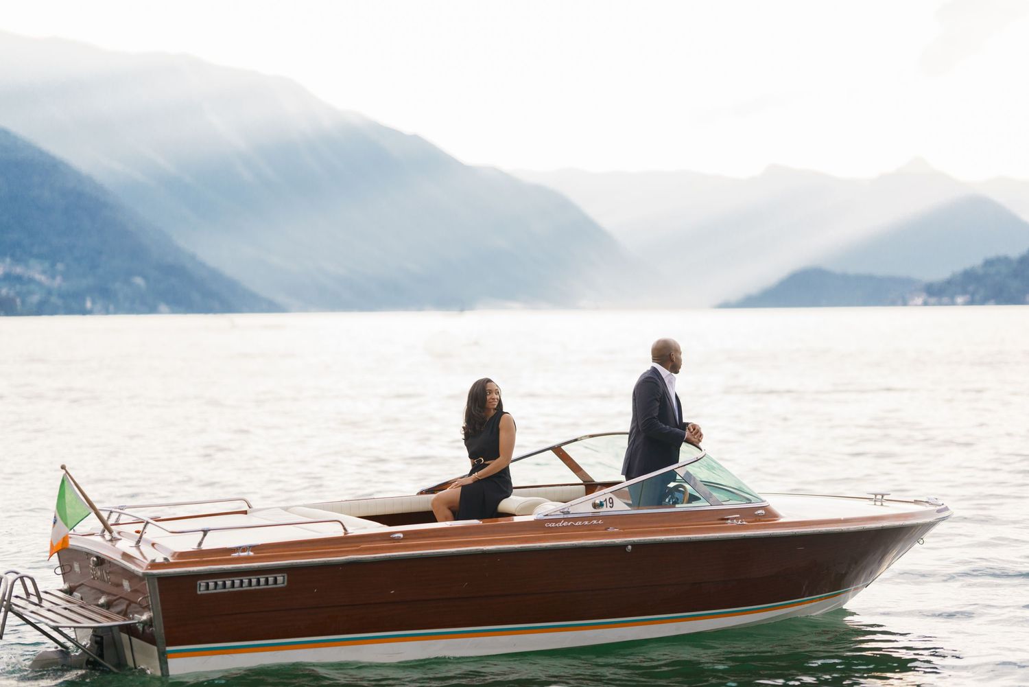 Two people cruise on a classic wooden boat across a misty mountain lake on a calm day.