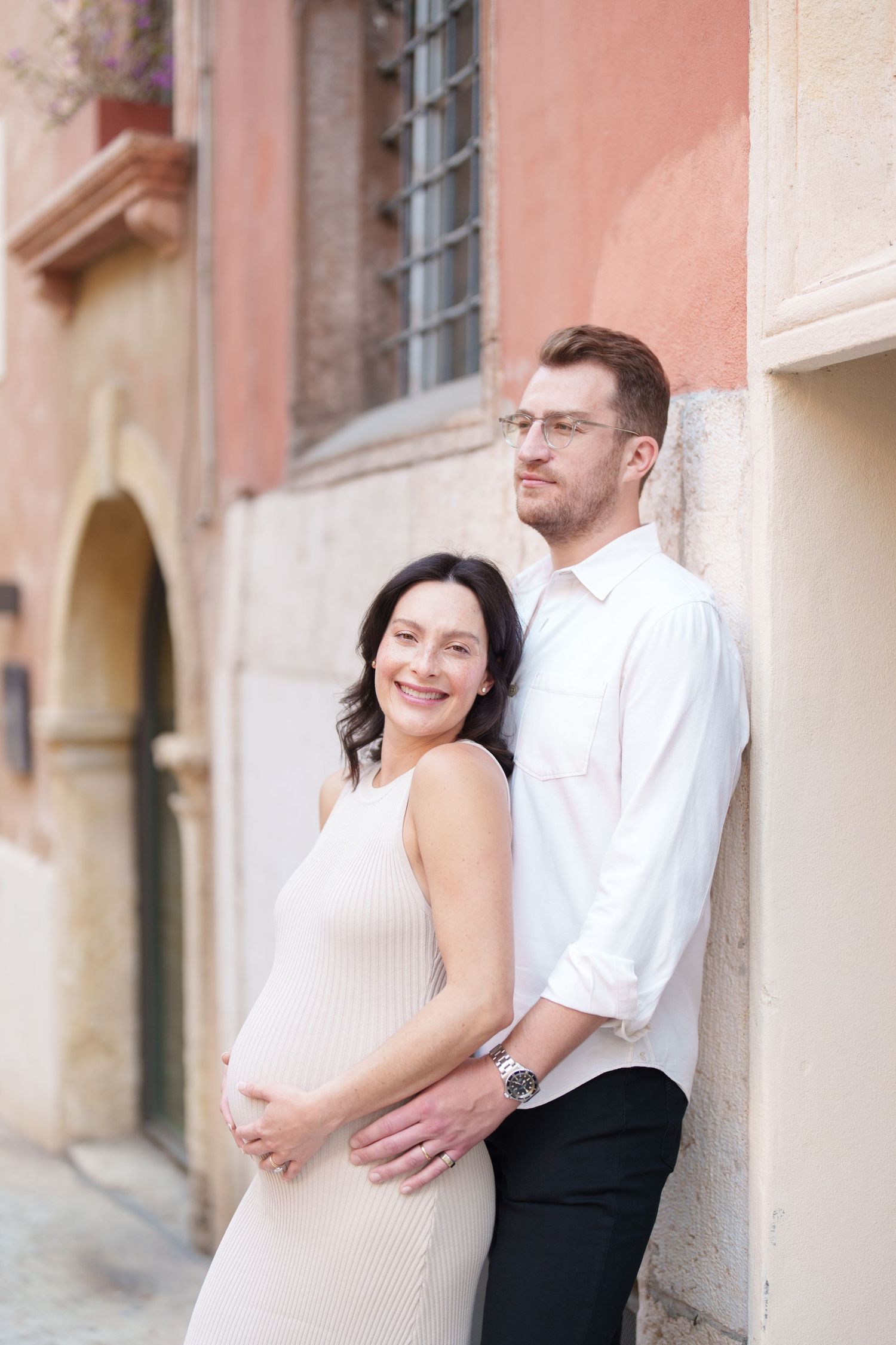 A couple poses together against a pink wall, with the woman wearing a cream dress showing her pregnant belly.