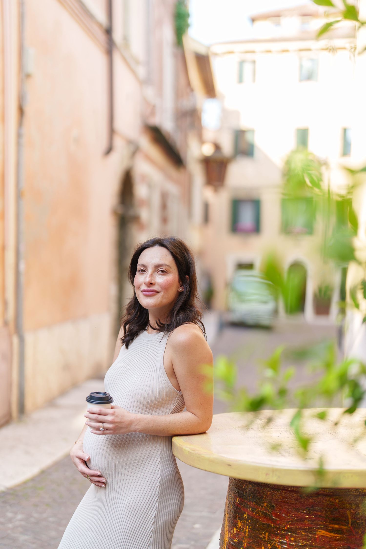 A person in a beige ribbed sleeveless top stands on a charming European street with stone buildings and greenery.