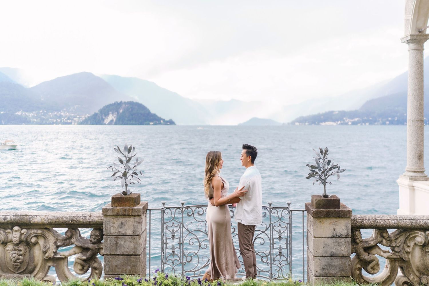 A couple stands on a scenic lakeside terrace overlooking mountains and water in Lake Como, Italy.