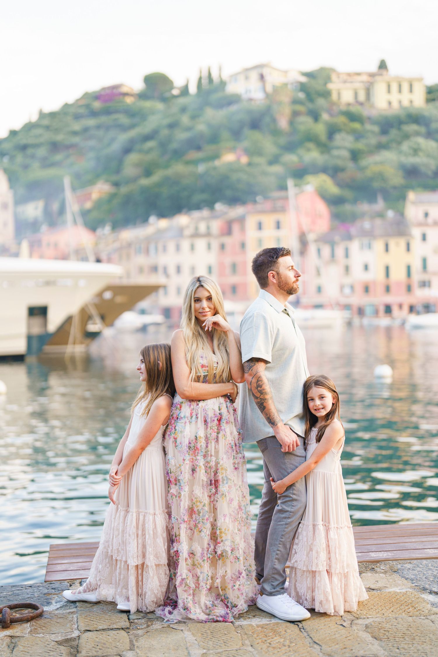 Family in coordinating pink and white outfits poses together along a scenic Mediterranean waterfront at sunset.