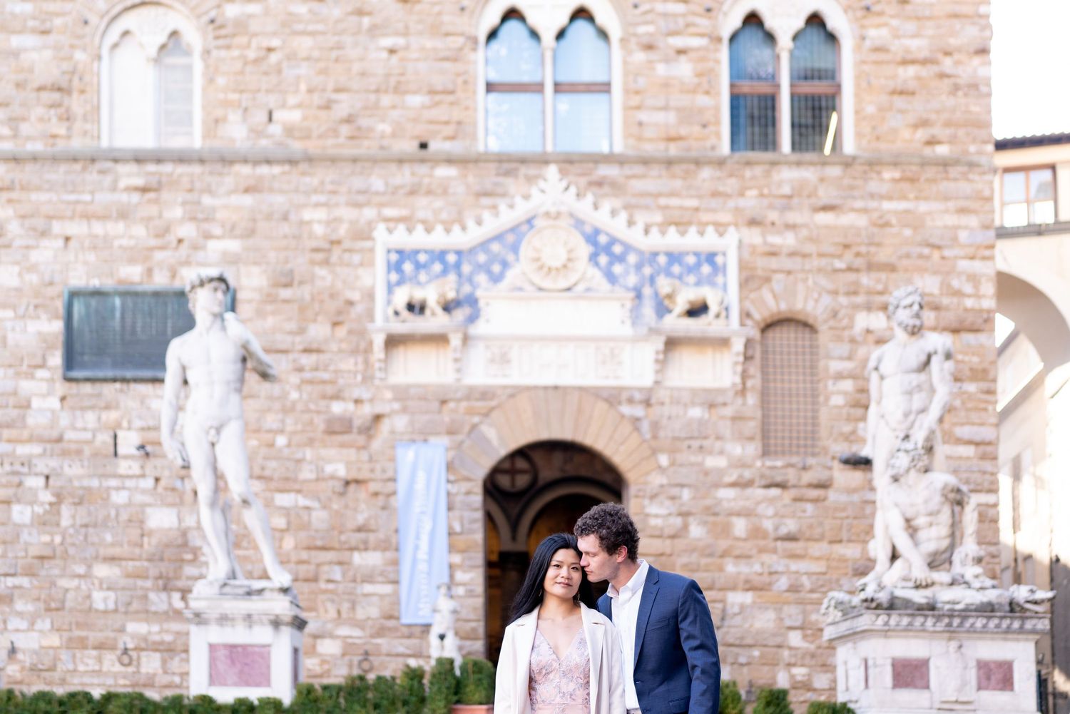 A romantic couple stands between classical statues outside an ornate stone building with Gothic windows in Florence, Italy.
