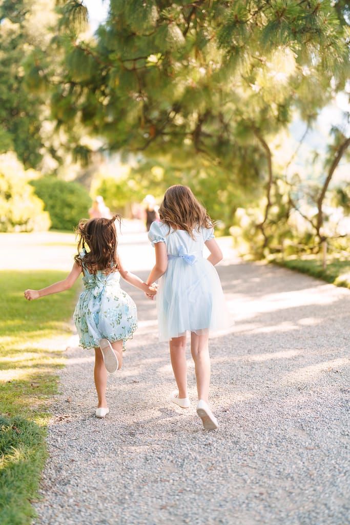 Two small children in light dresses walk hand in hand down a gravel path surrounded by trees.