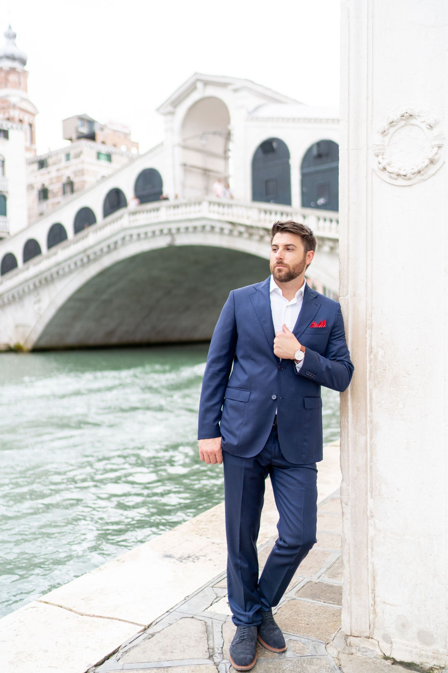 A person in a navy blue suit poses along the canals of Venice with the iconic Rialto Bridge visible in the background.