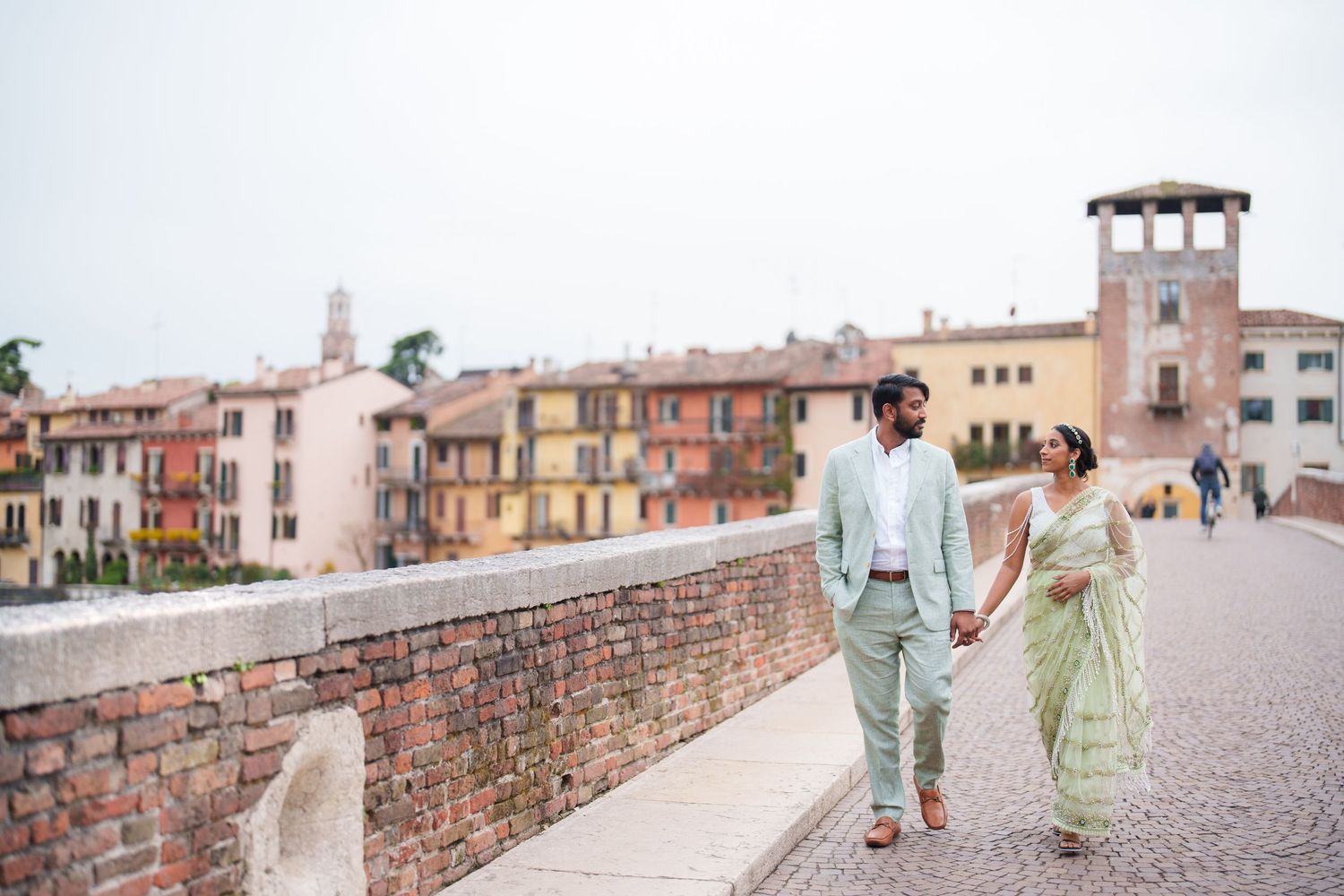 A couple walks hand in hand along a scenic stone wall overlooking an Italian village at sunset.
