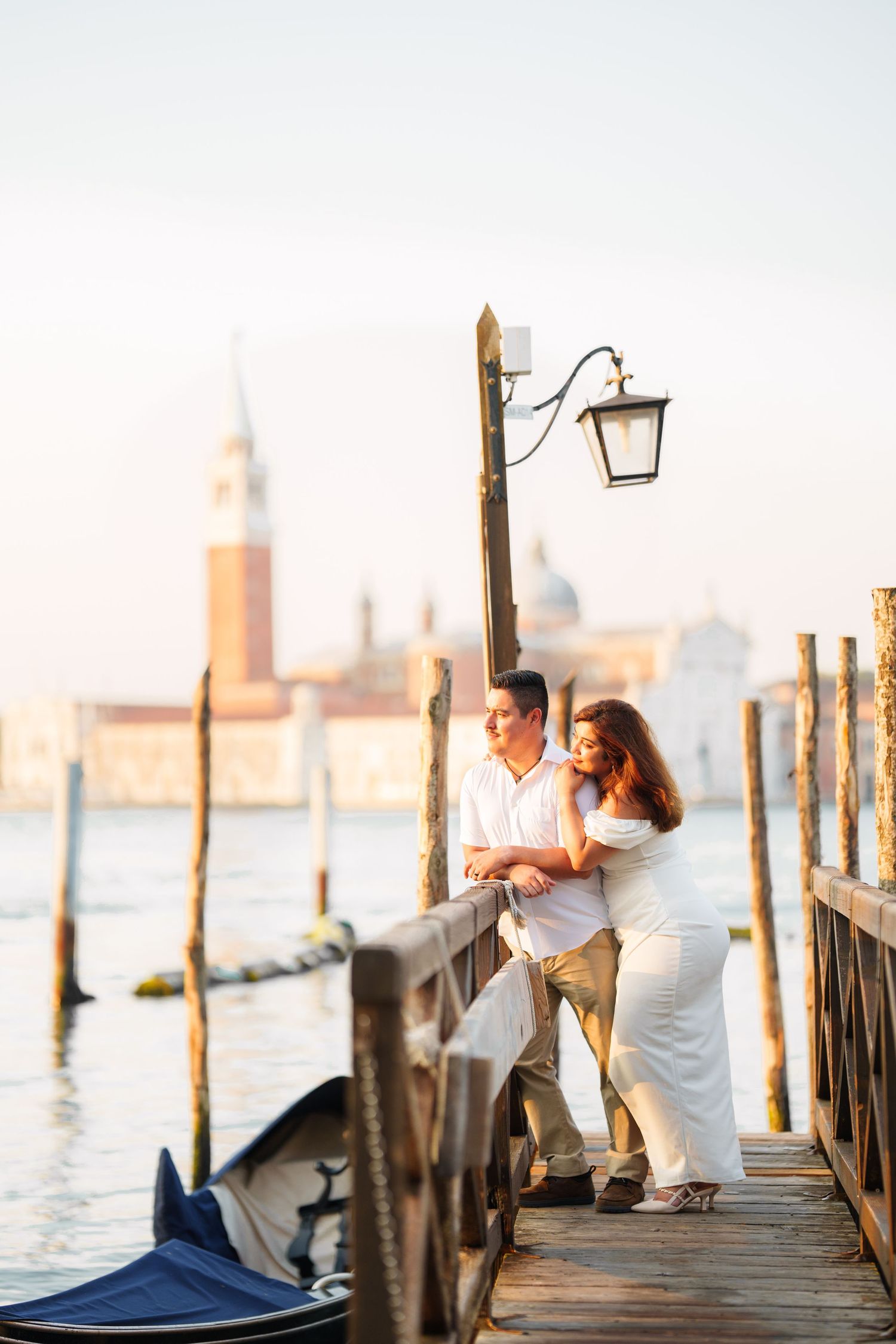 Romantic couple embraces on wooden pier at sunset with Venice cityscape in background across lagoon.