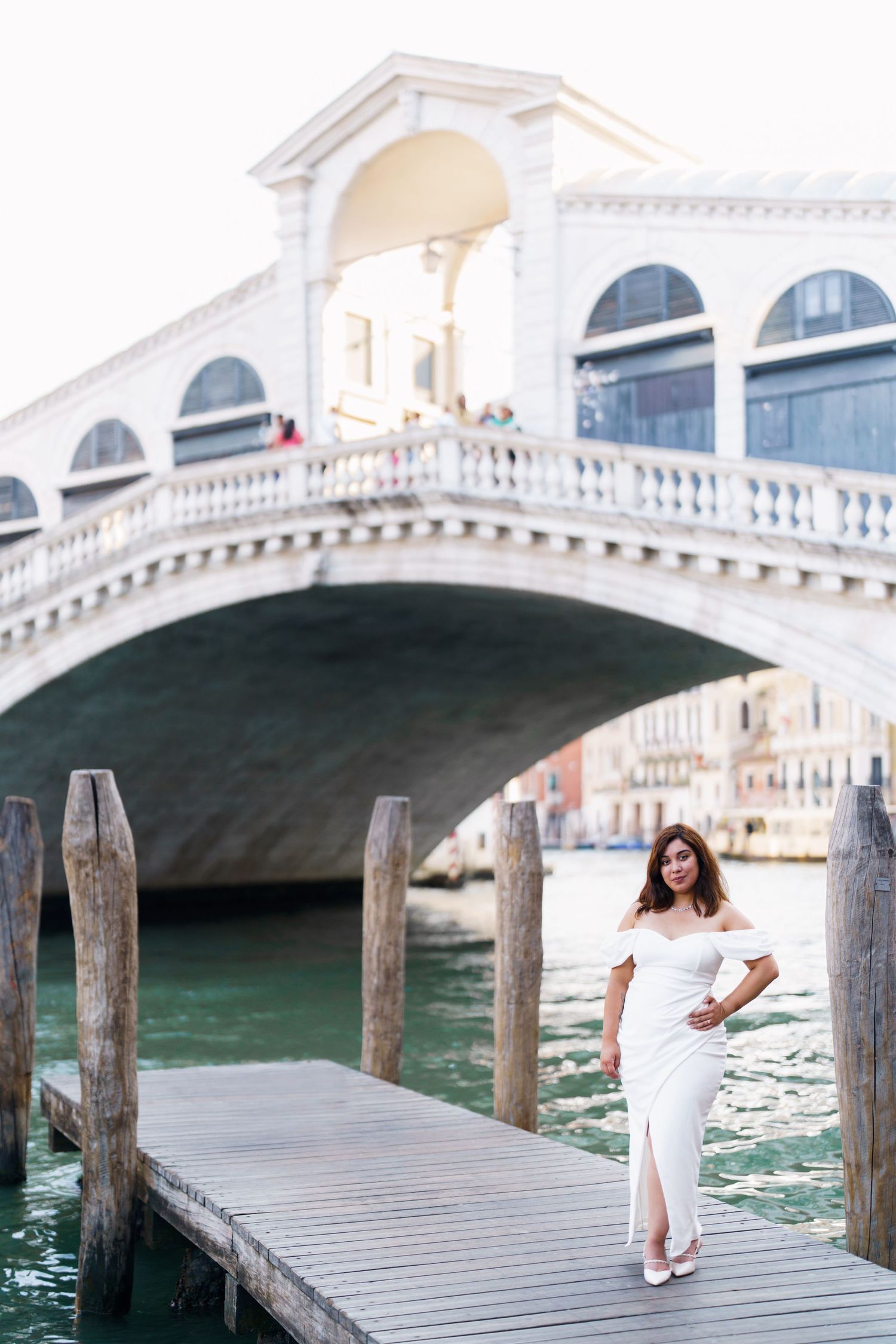 photographer venice Woman in white dress poses on wooden dock by Venice's Rialto Bridge and canal waterfront.
