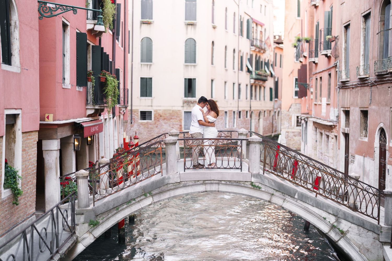 A romantic scene on a small arched bridge over a Venice canal with pink buildings and traditional architecture.